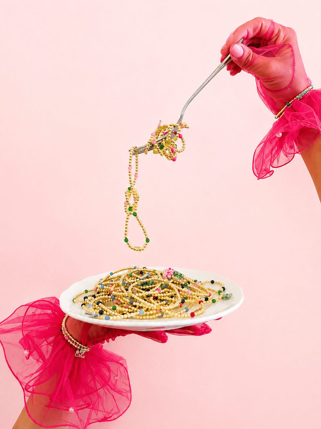 Person wearing pink gloves and ruffled sleeves holds a fork with jewelry over a plate of jewelry against a pink background.