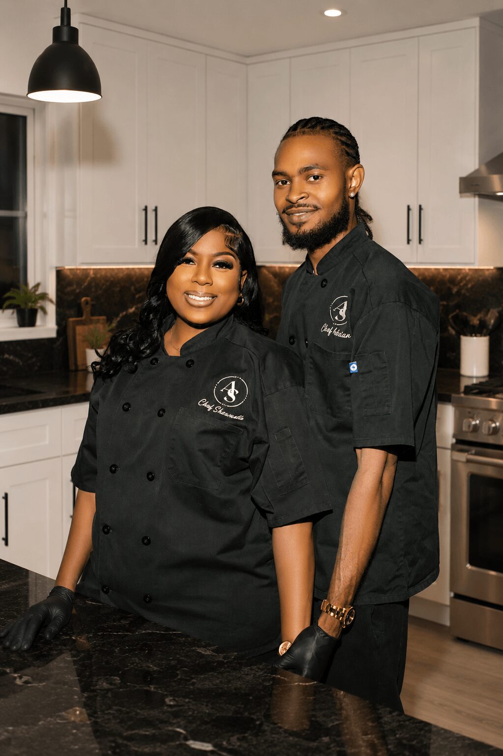 Two chefs in black uniforms stand in a modern kitchen, smiling at the camera.