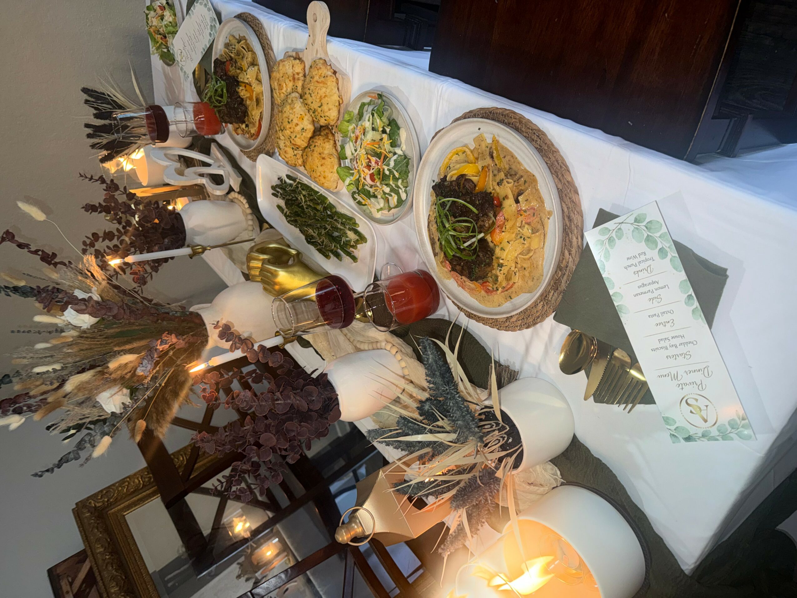 Table with various dishes, drinks, and a menu, decorated with dried flowers and candles, viewed from the side.