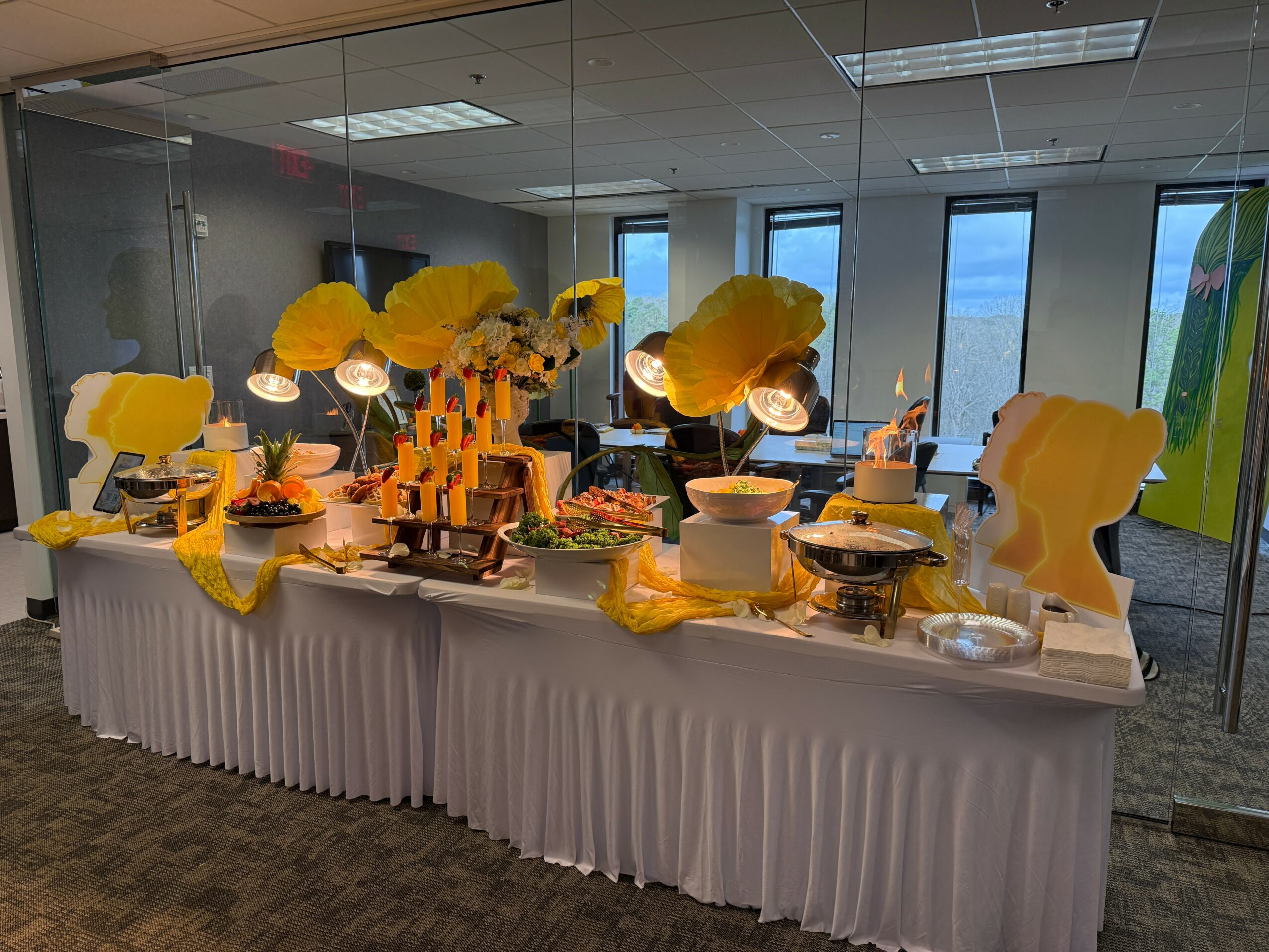 Long buffet table with yellow decorations, food, and serving dishes in a room with large windows and ceiling lights.