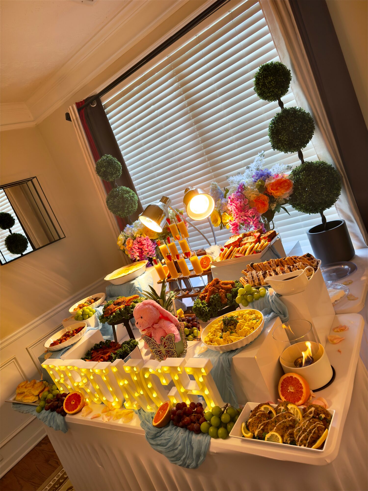 Table with food and decorations, including topiary trees, flowers, and a lit sign, viewed from the side with window blinds in background.