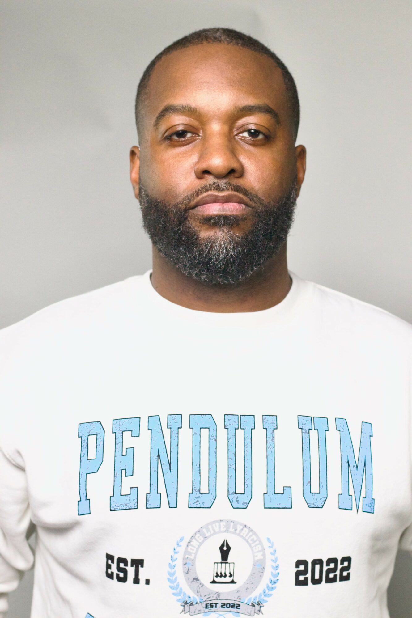 Man with beard and short hair wearing a white sweatshirt with blue text, neutral expression, plain background.