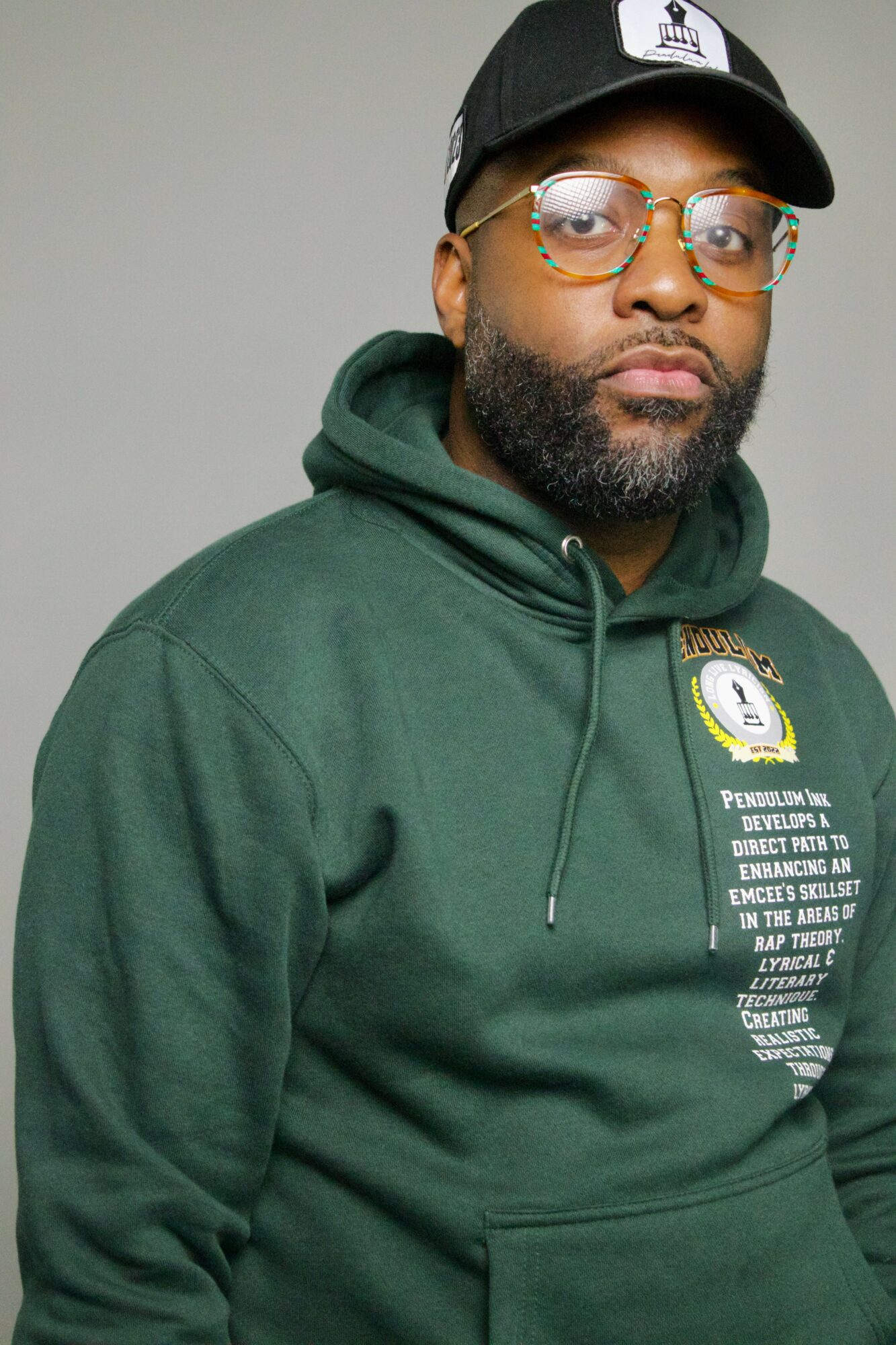 Man wearing glasses, black cap, green hoodie with text, against a plain background.