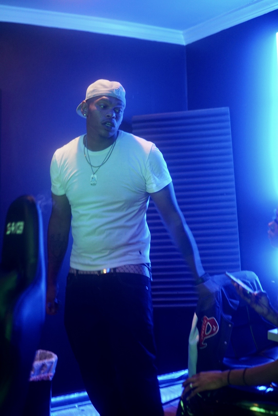 Man wearing a white t-shirt, cap, and necklace in a room with blue lighting, standing near a person sitting down.