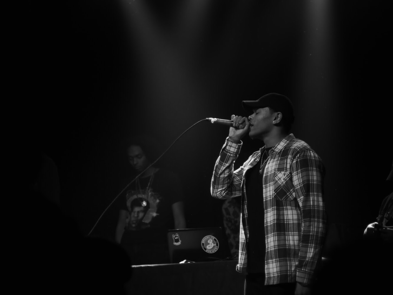 Person singing into a microphone on stage with a DJ in the background, black and white image.