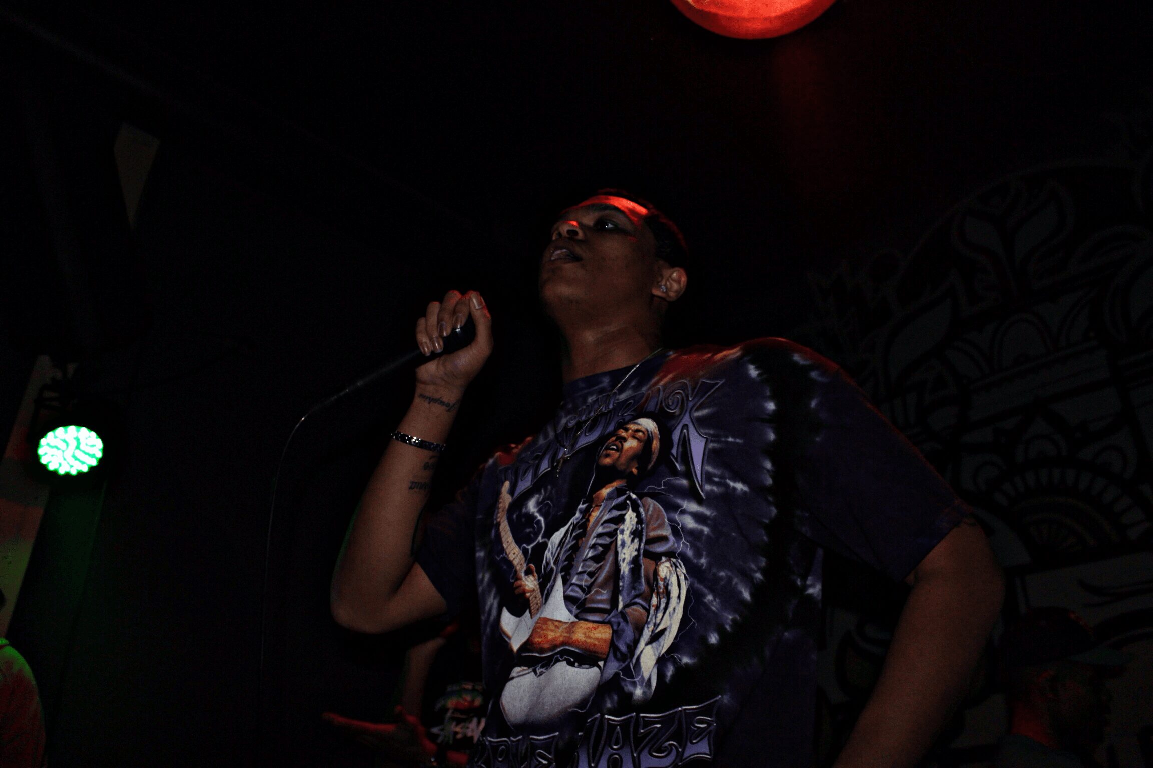 Person singing into a microphone on stage with dark background and colorful lighting.