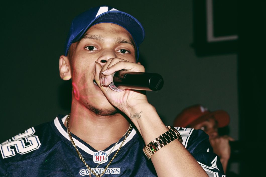 Young man wearing a cap and sports jersey, holding a microphone close to his mouth, performing on stage.