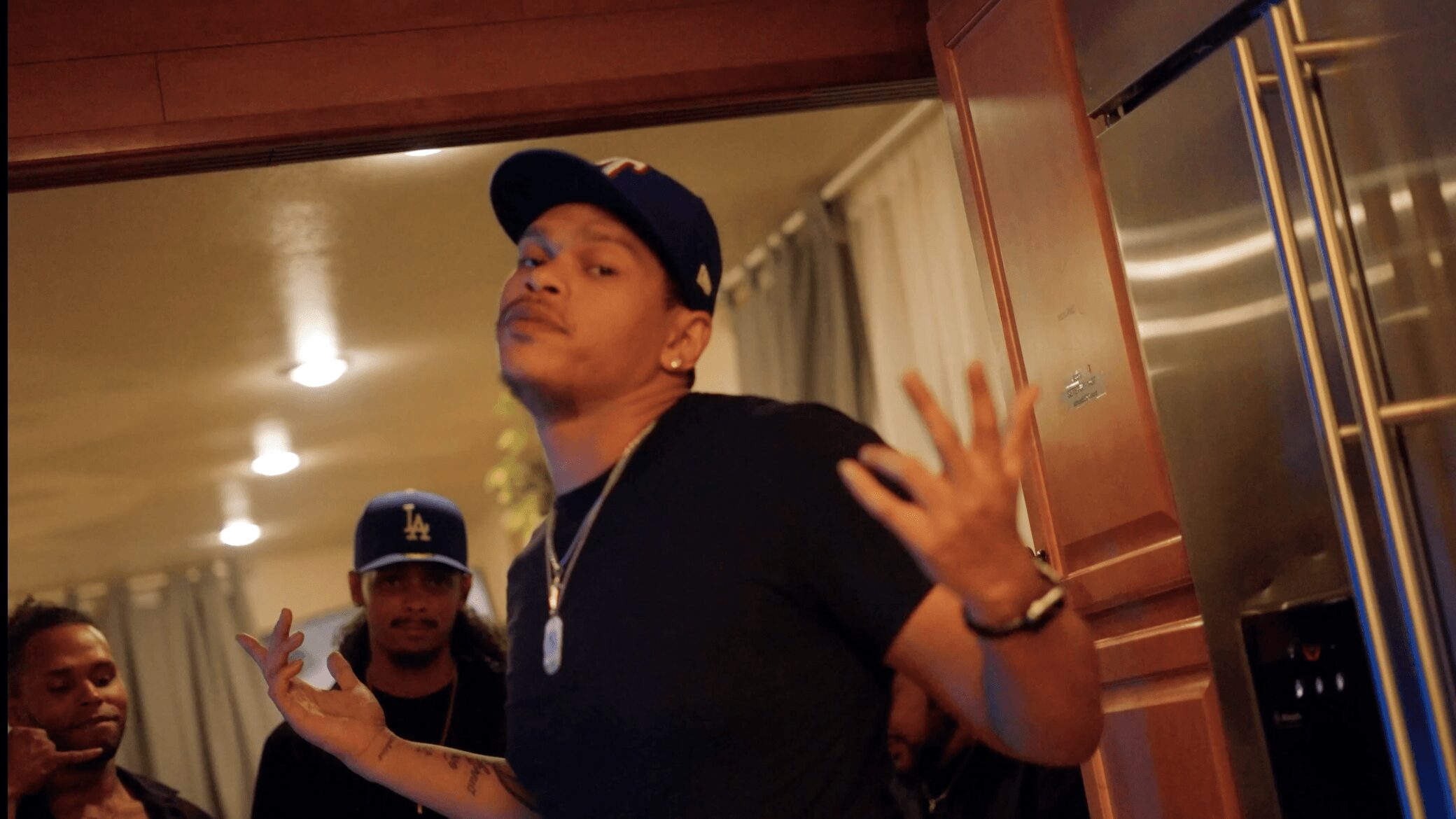 Person in black shirt and cap gesturing with hands, others in background, indoor setting.