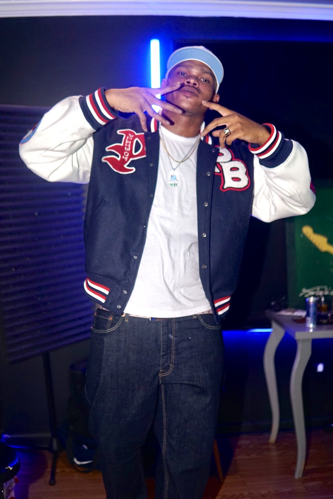 Young man wearing a baseball cap and varsity jacket making hand gestures indoors.