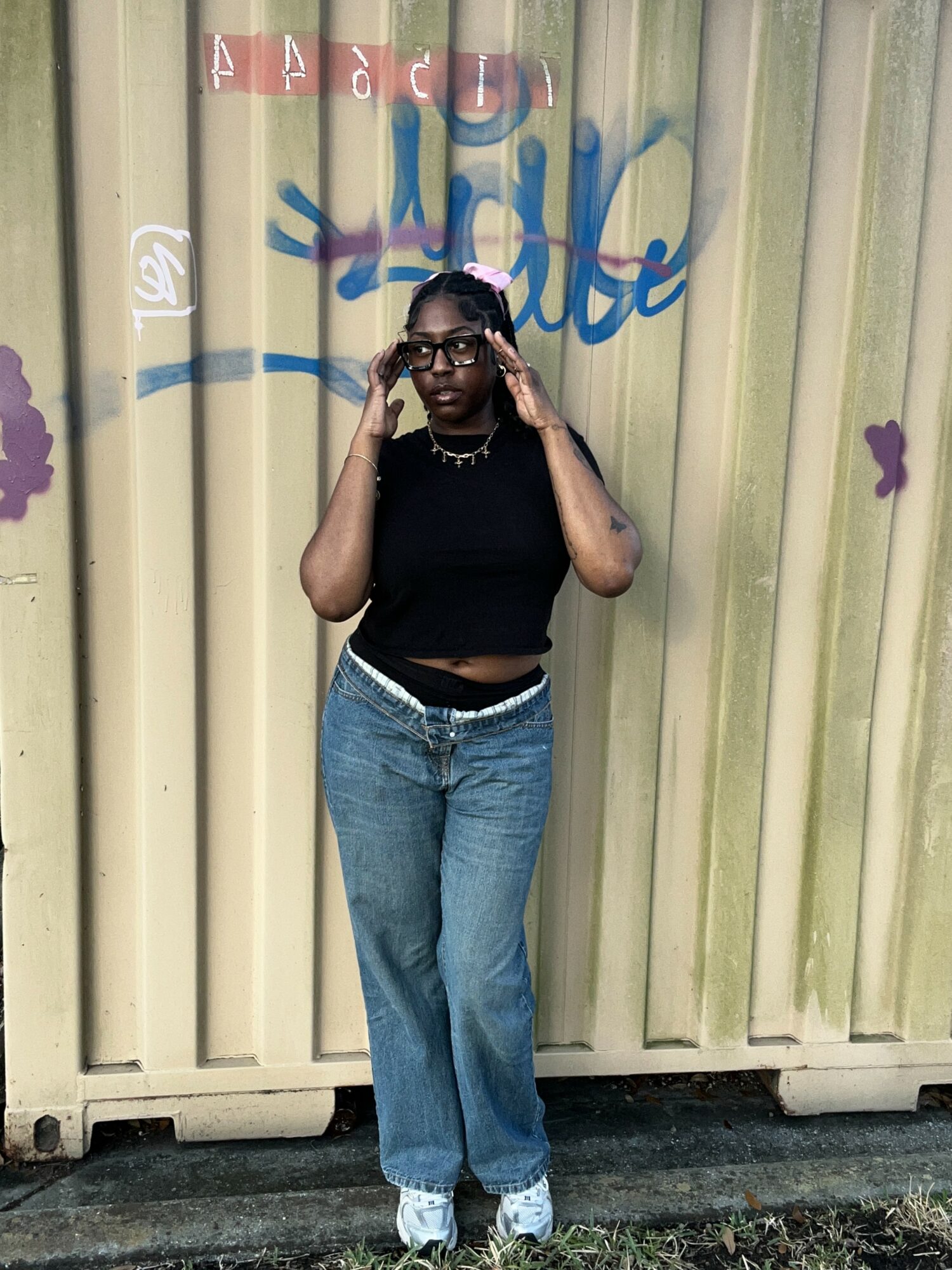 Young woman with glasses and curly hair stands in front of a graffiti-covered wall, adjusting her glasses, wearing a black crop top and jeans.