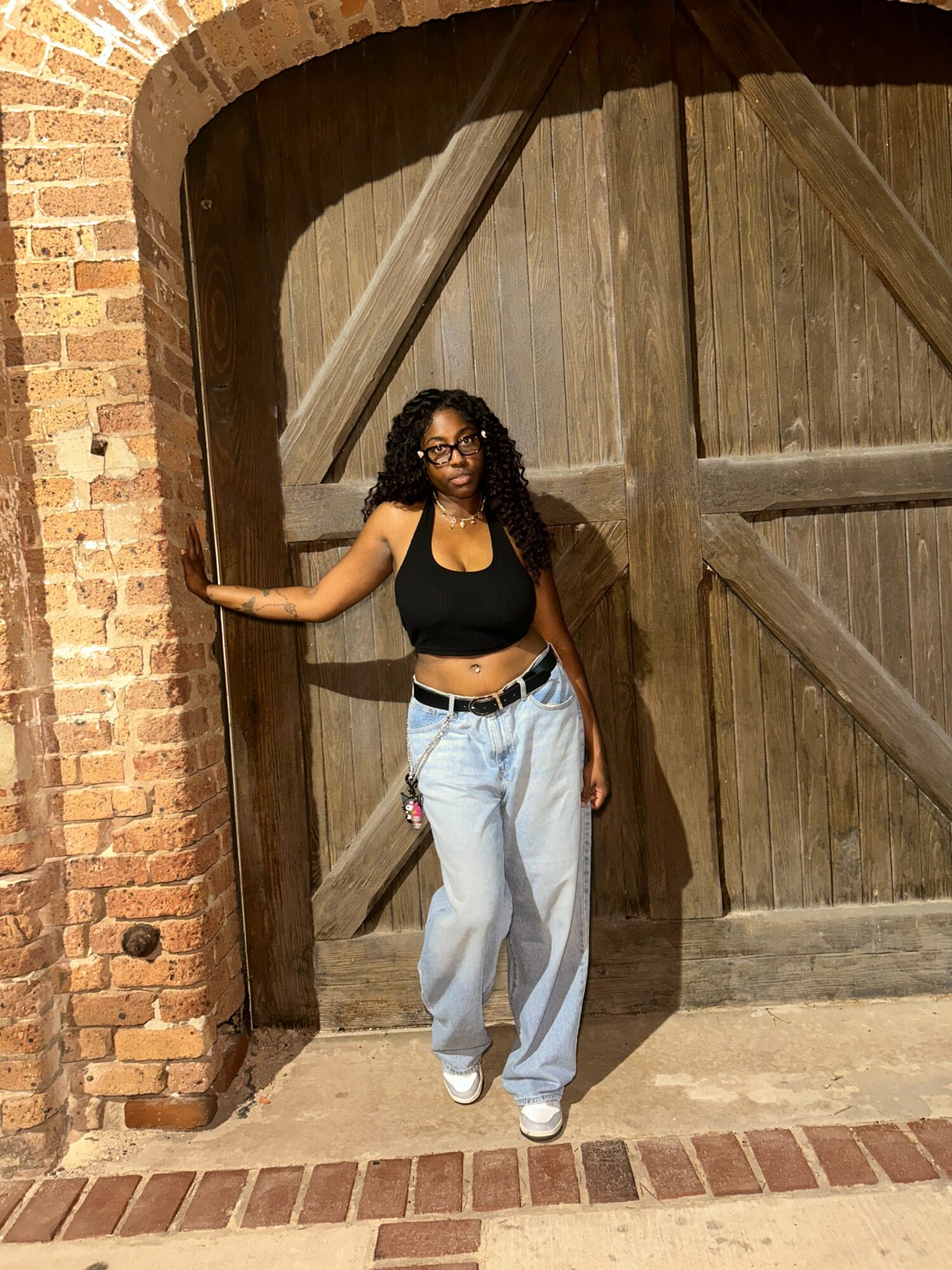 Woman with curly hair and glasses stands in front of a large wooden door, holding the brick wall with one hand.