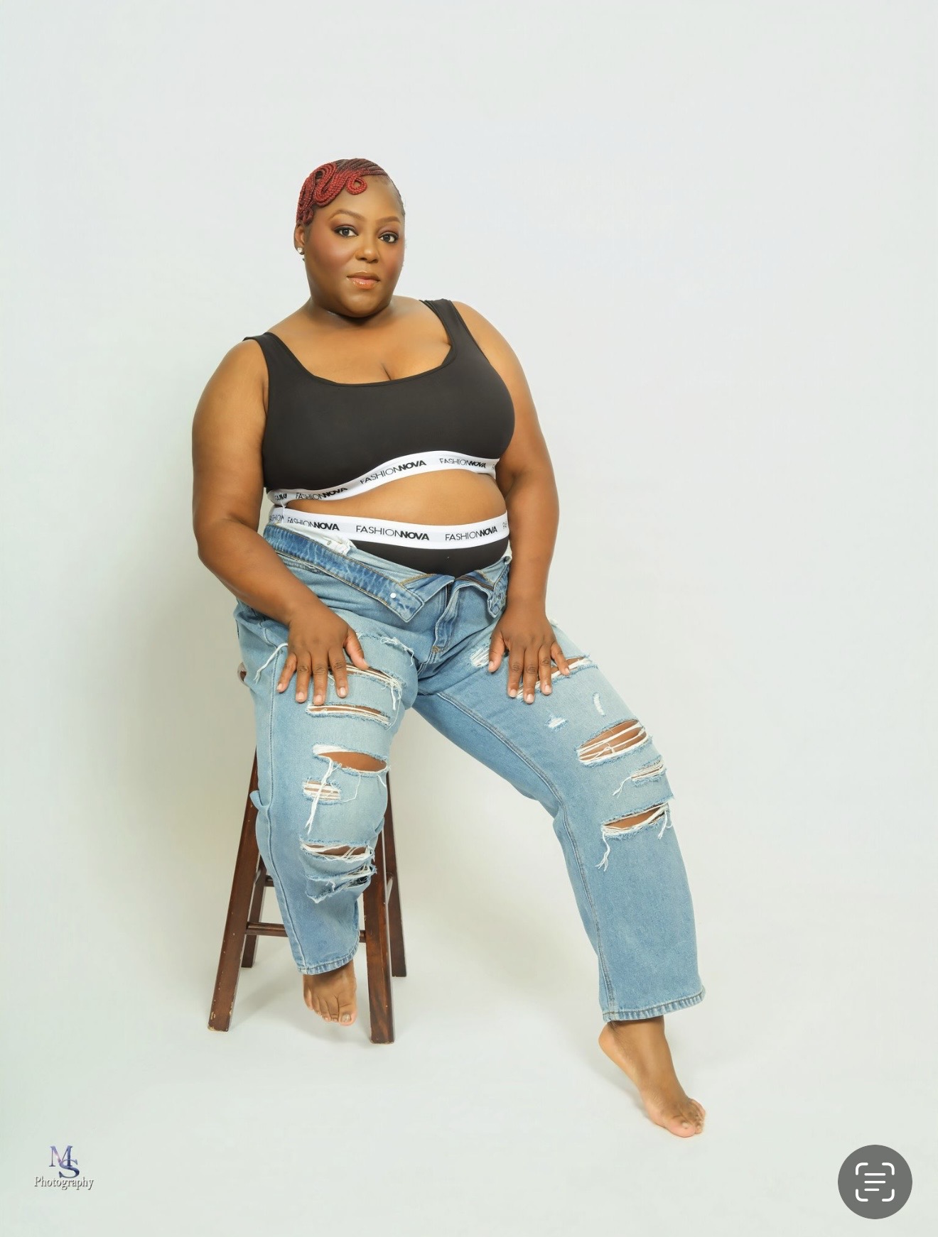 Woman with red hair sitting on a stool, wearing a black top and ripped jeans, against a plain light background.