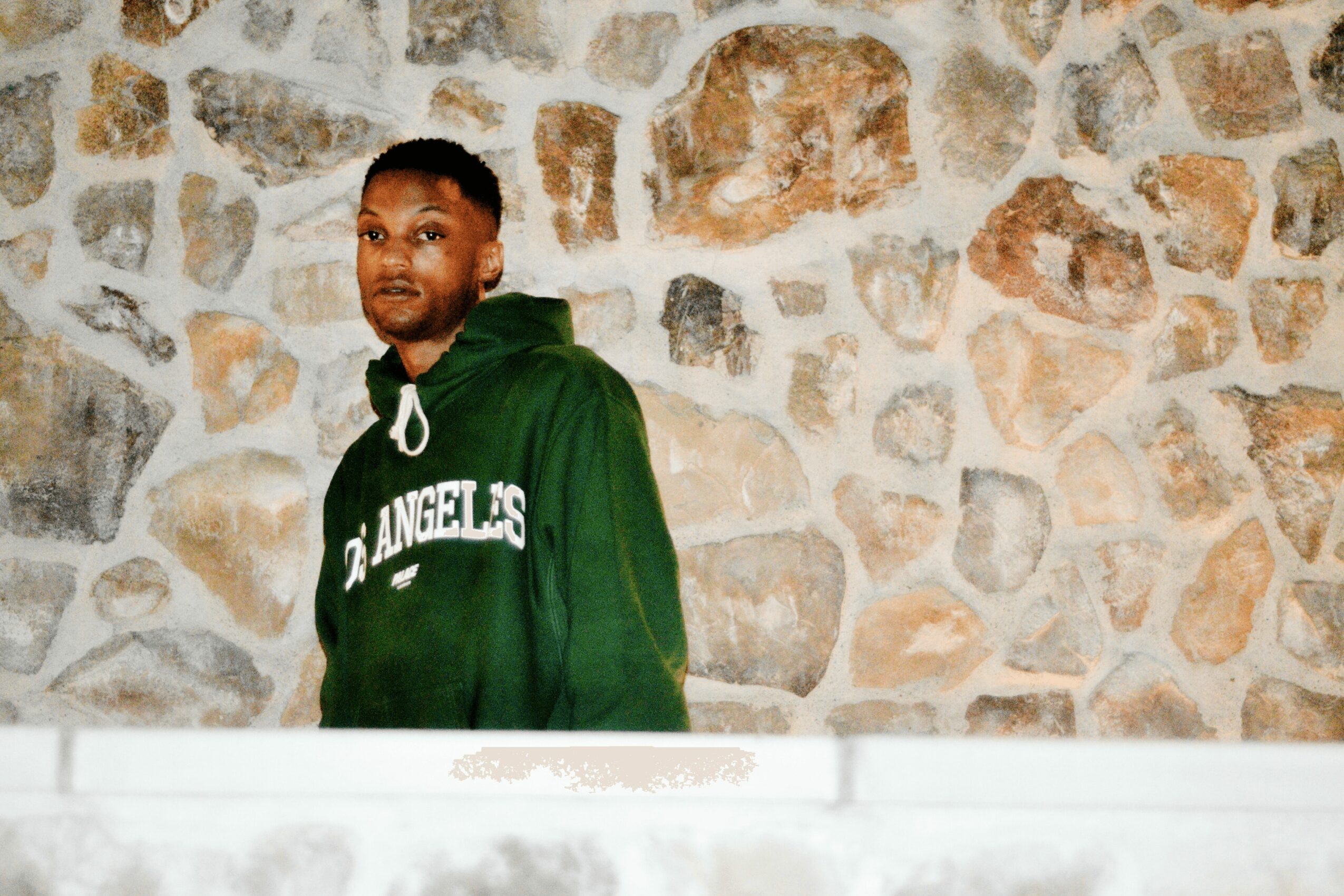 Young man in green hoodie standing against a stone wall, looking to the side, smiling slightly.