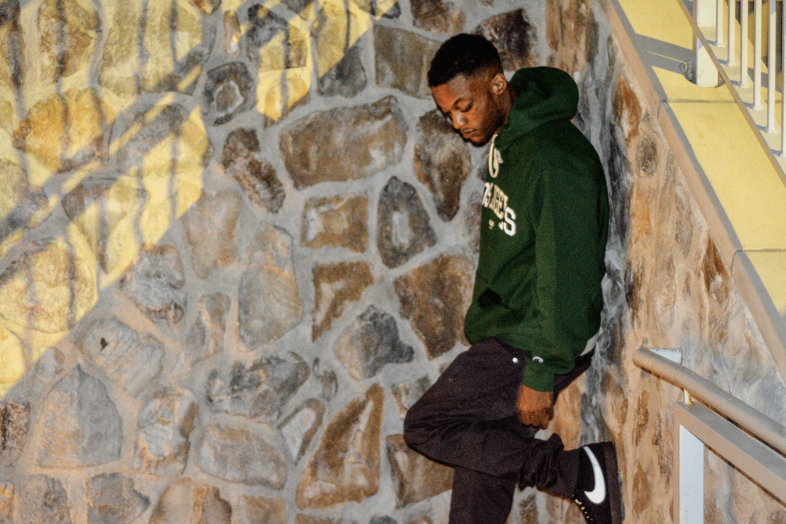 Young man leaning against a stone wall, wearing a green hoodie and black pants, looking down.