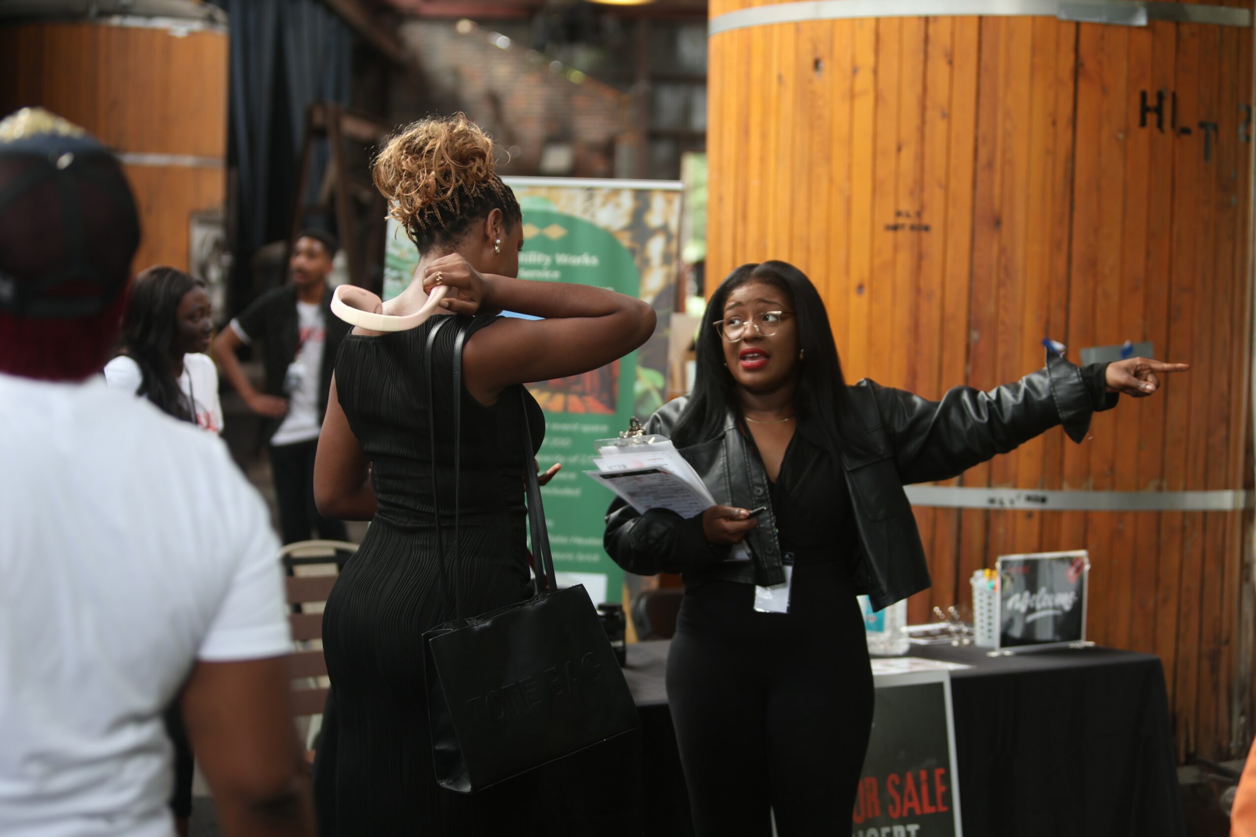 Two women engaged in conversation outdoors, one gesturing with her arm, others in the background, wooden structures nearby.