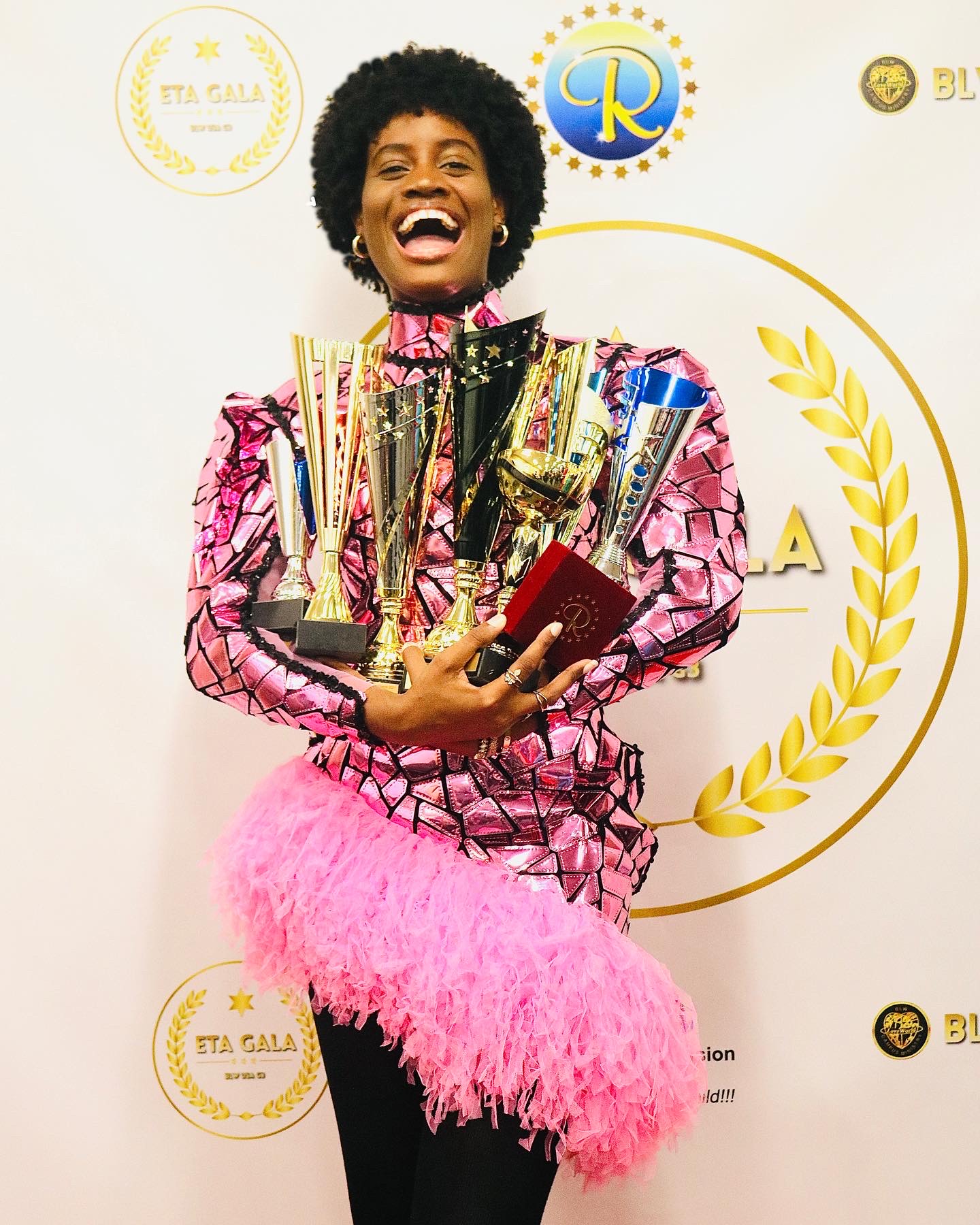 Woman with curly hair smiling, holding multiple trophies and a red book, wearing a pink feathered skirt and patterned top.