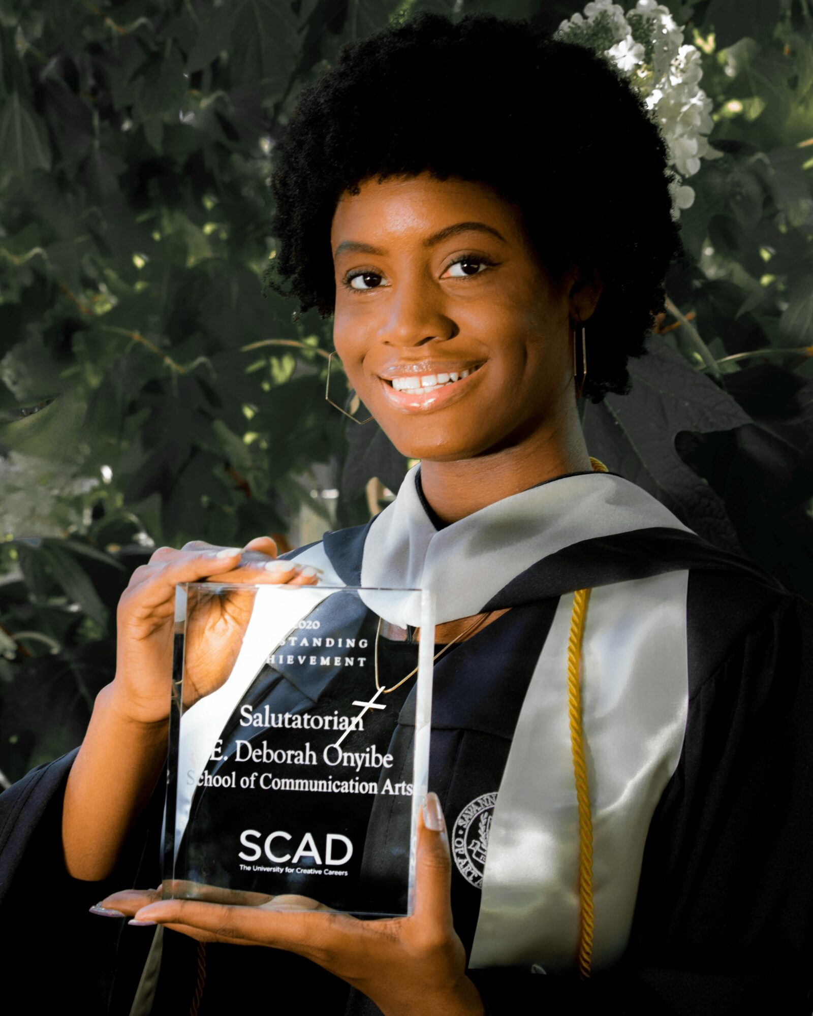 Young woman in graduation gown holding a clear award plaque, smiling, with green foliage background.