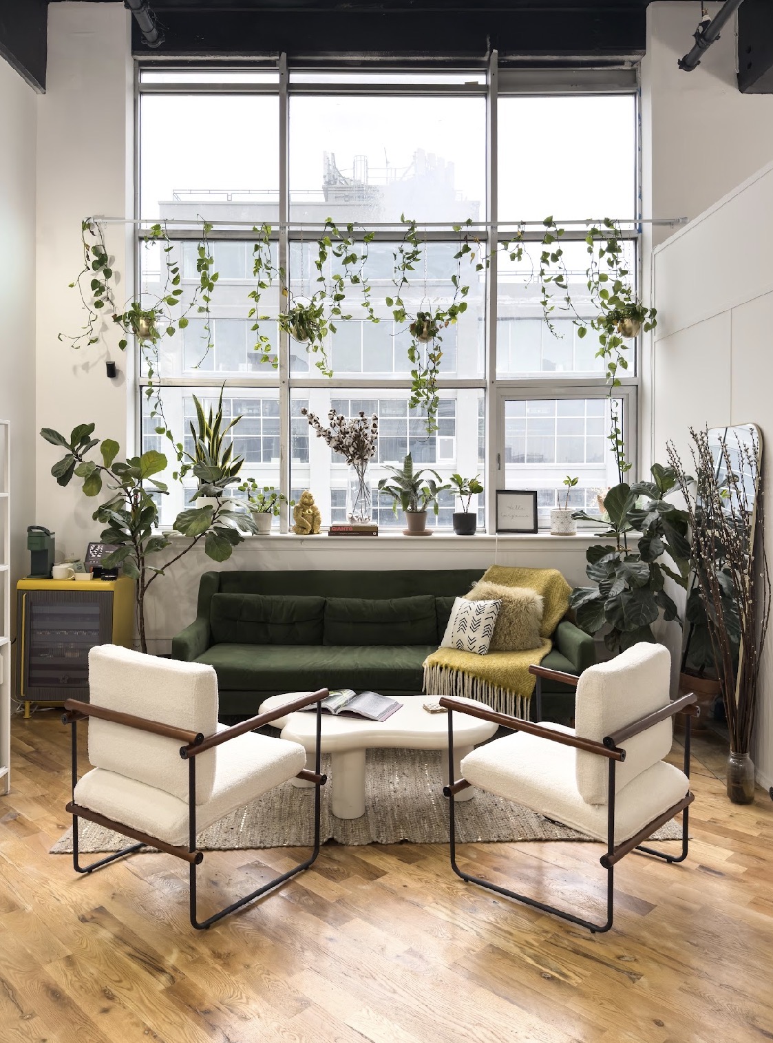 Interior with large window, green sofa, two white armchairs, plants, and wooden floor.