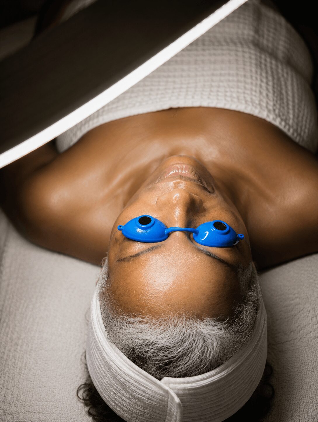 Elderly person lying down with eyes closed, wearing goggles and a headband, covered with a towel, in a spa or treatment setting.