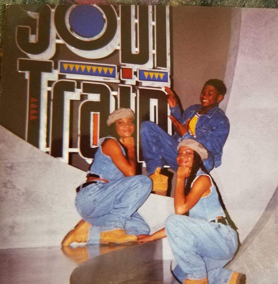Three people sitting and smiling in front of a colorful sign, with one person raising an arm. All are wearing denim outfits and hats.