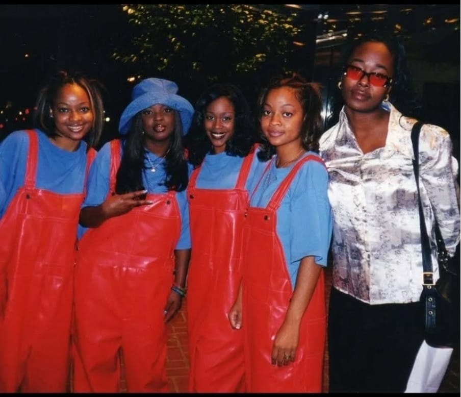 Five women standing outdoors at night, four wearing blue shirts and red overalls, one in a patterned blouse with glasses, smiling.