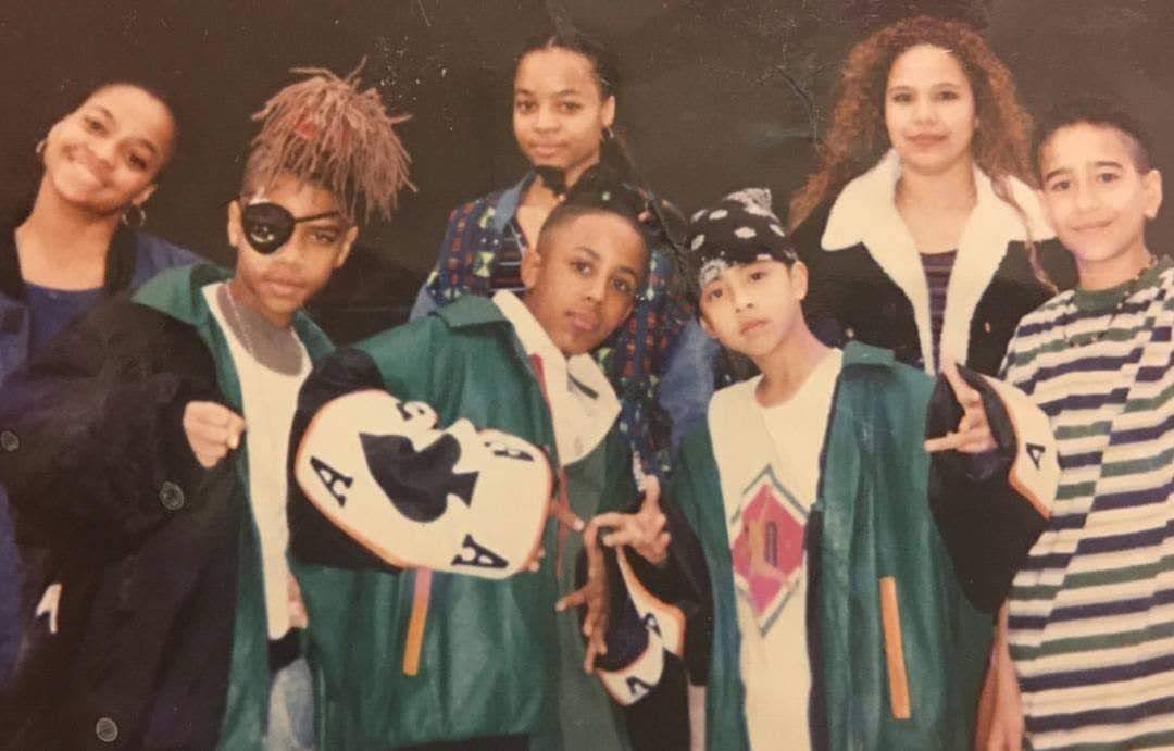 Group of seven children and teenagers posing together, some wearing green jackets, in a dark background.
