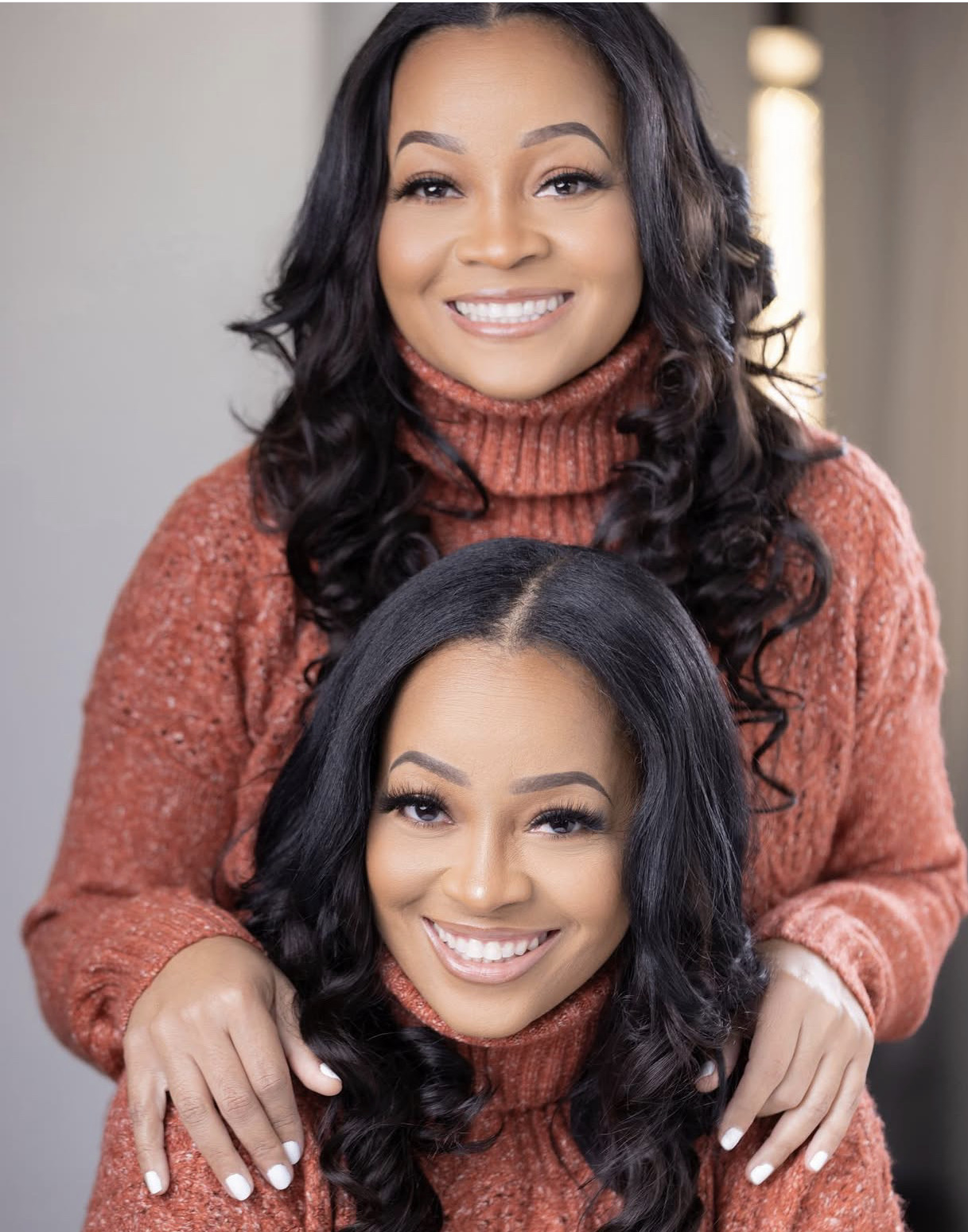 Two women with long dark hair smiling, wearing matching rust-colored sweaters, one standing behind the other.