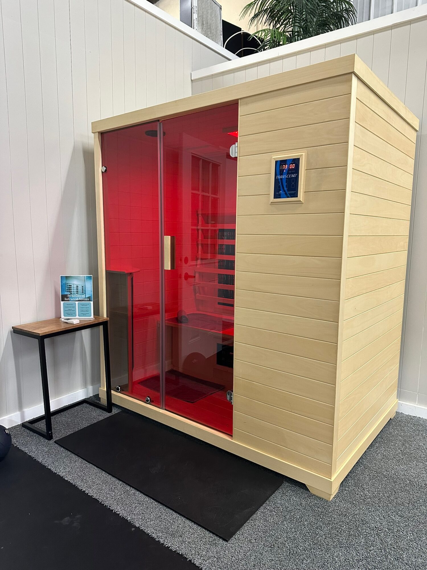 Red glass sauna with wooden exterior, small table with brochures outside, black mat in front, white wall background.