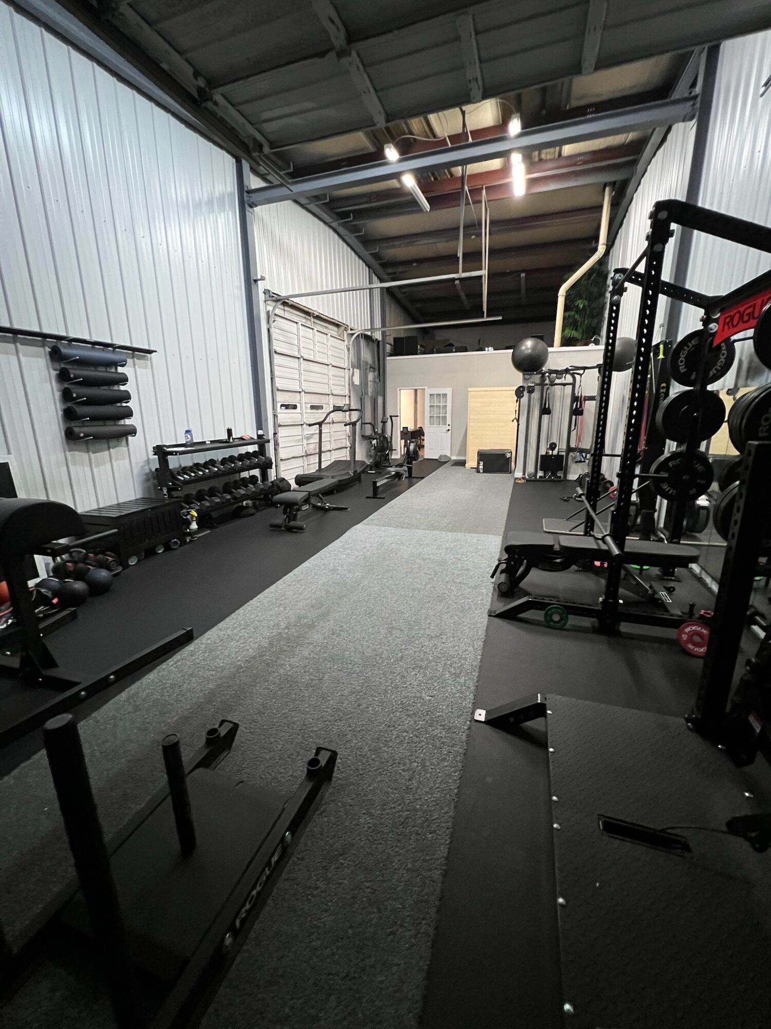 Gym interior with workout equipment, benches, and weights along walls, with a central carpeted area, metal walls, and ceiling.