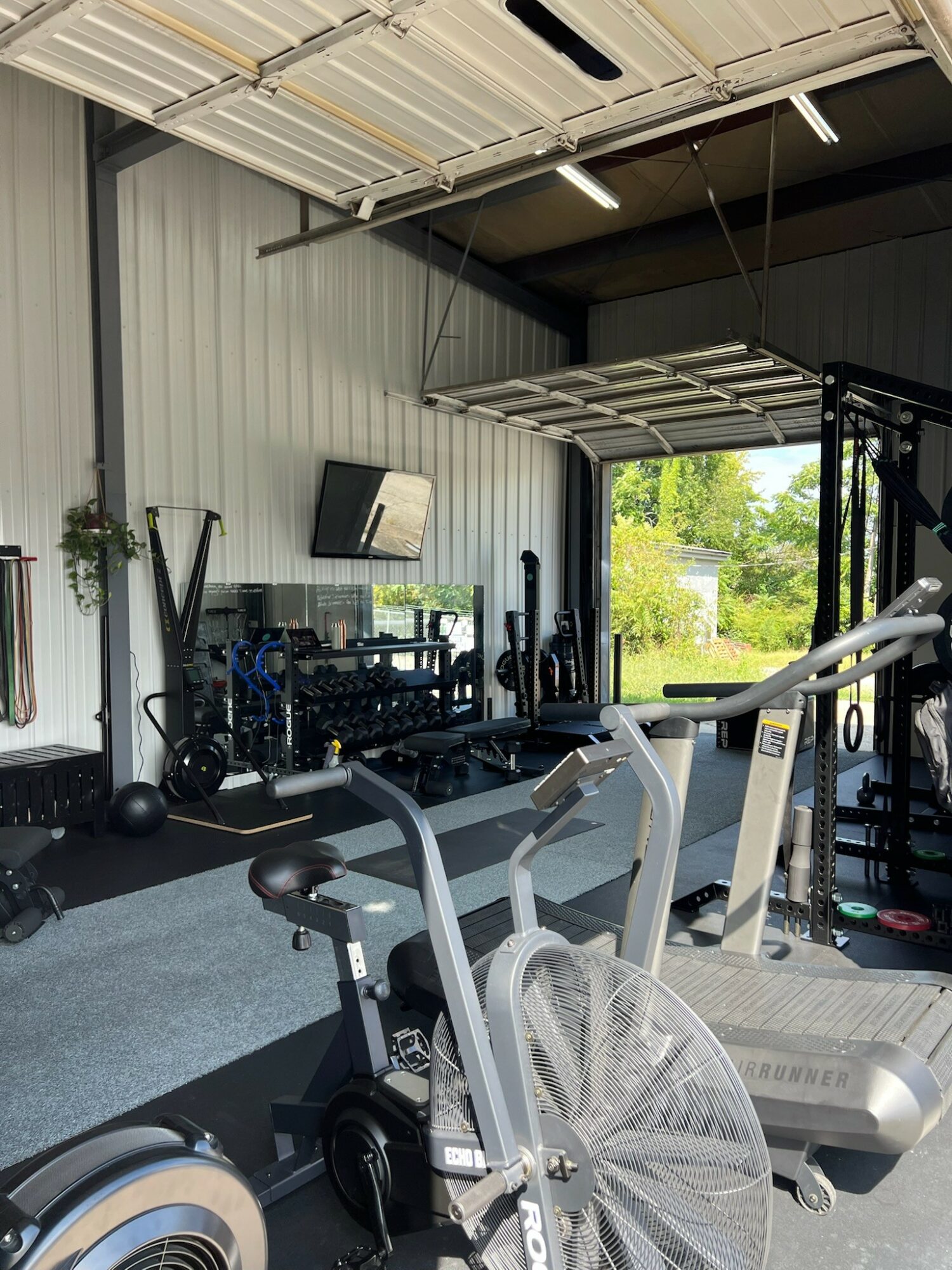 Gym interior with cardio equipment, weights, and a wall-mounted TV, near large open garage door showing outdoor greenery.