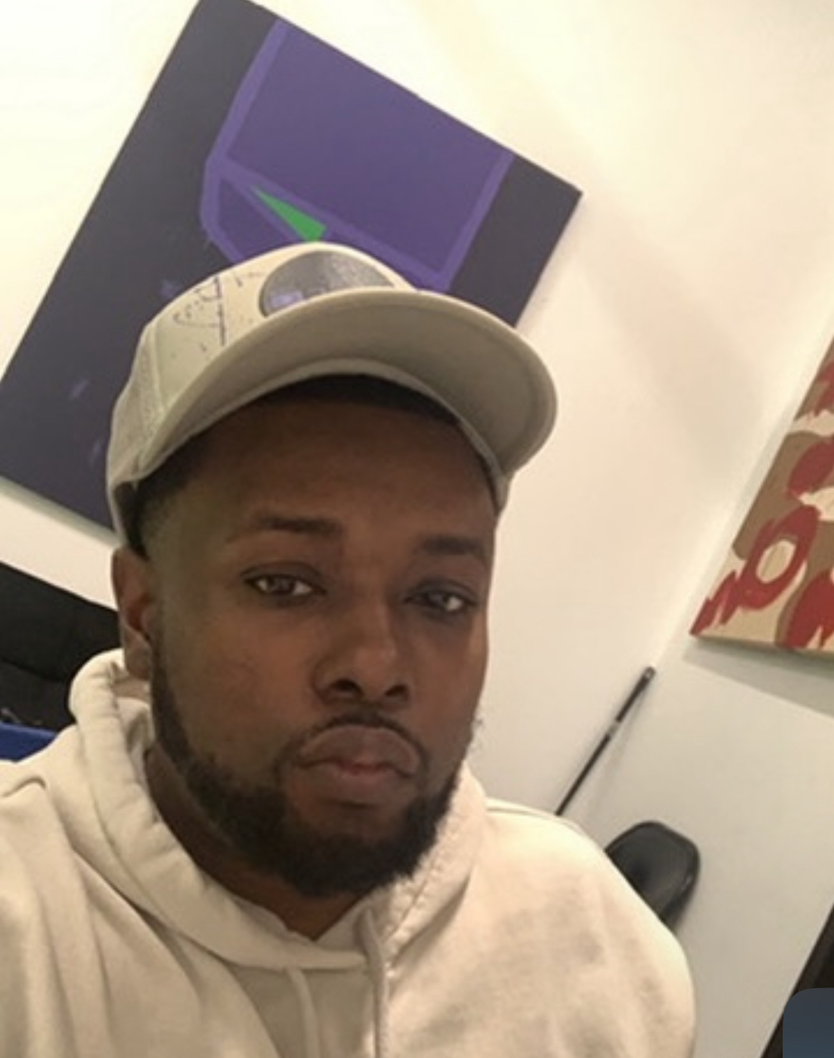 A man wearing a baseball cap and hoodie taking a selfie indoors, with artwork on the wall behind him.