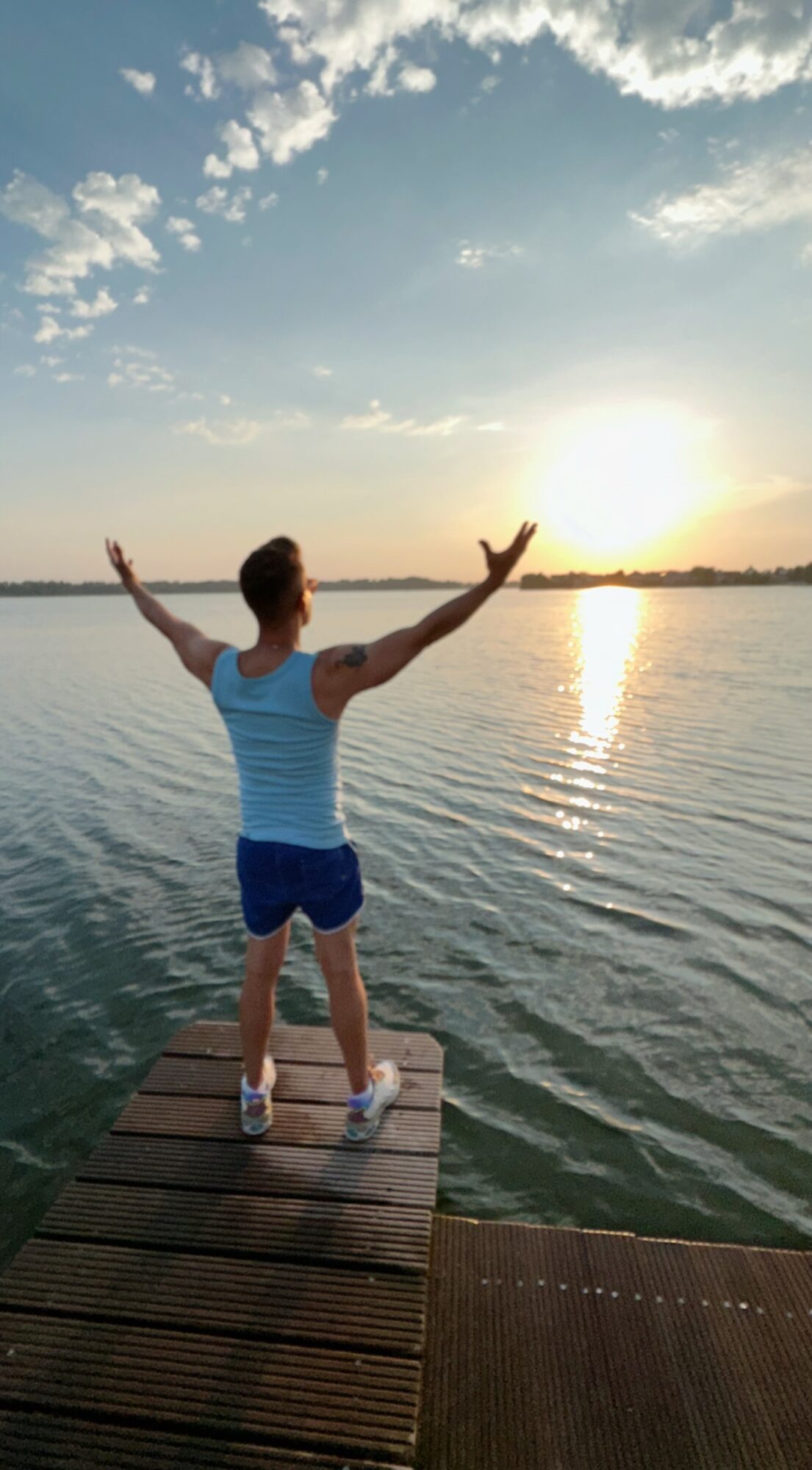 Person standing on a dock with arms raised, facing a body of water during sunset, sky partly cloudy.