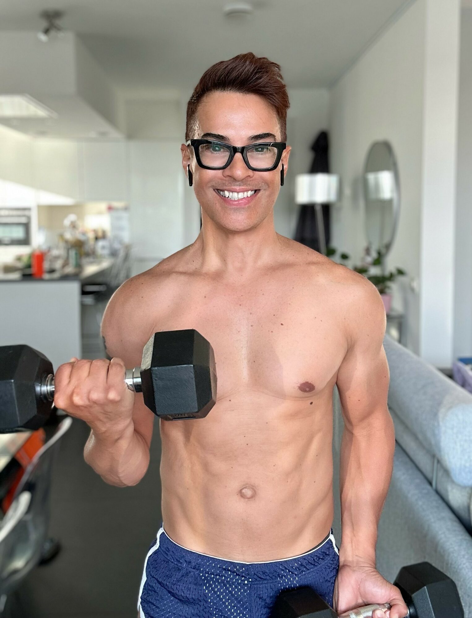 Person with short hair, glasses, smiling, holding a dumbbell, shirtless, in a modern kitchen and living space.