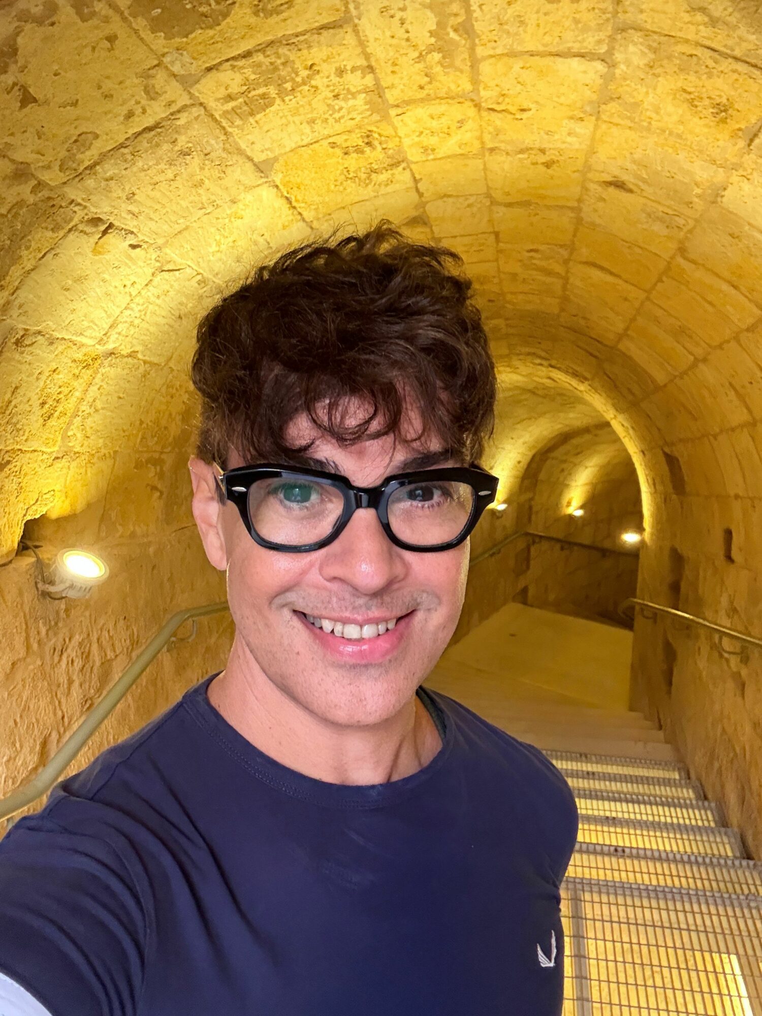 Person with short curly hair and glasses smiling in a tunnel with yellow brick walls.
