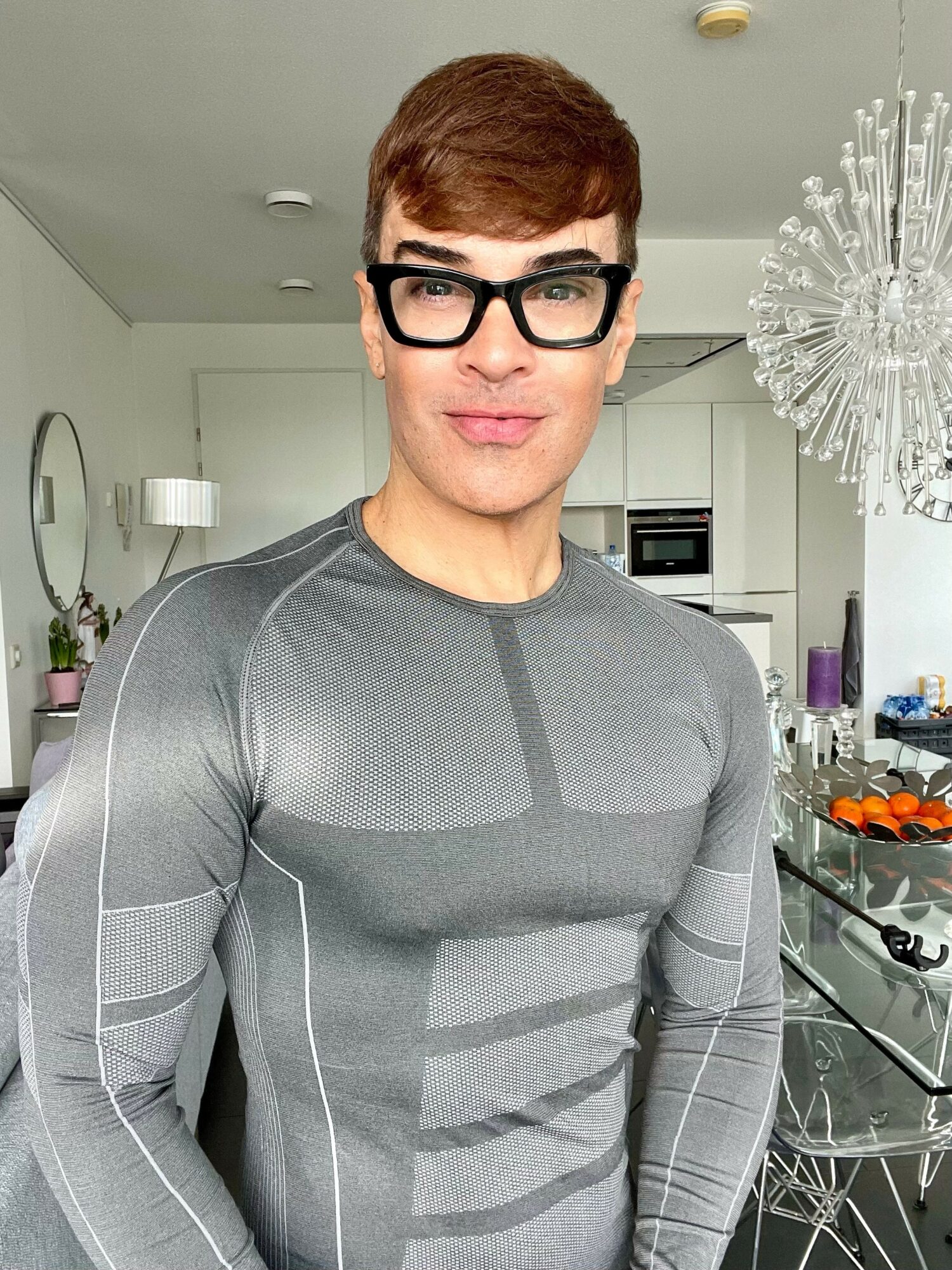 Person with short brown hair and black glasses wearing a gray long-sleeve shirt in a modern kitchen.