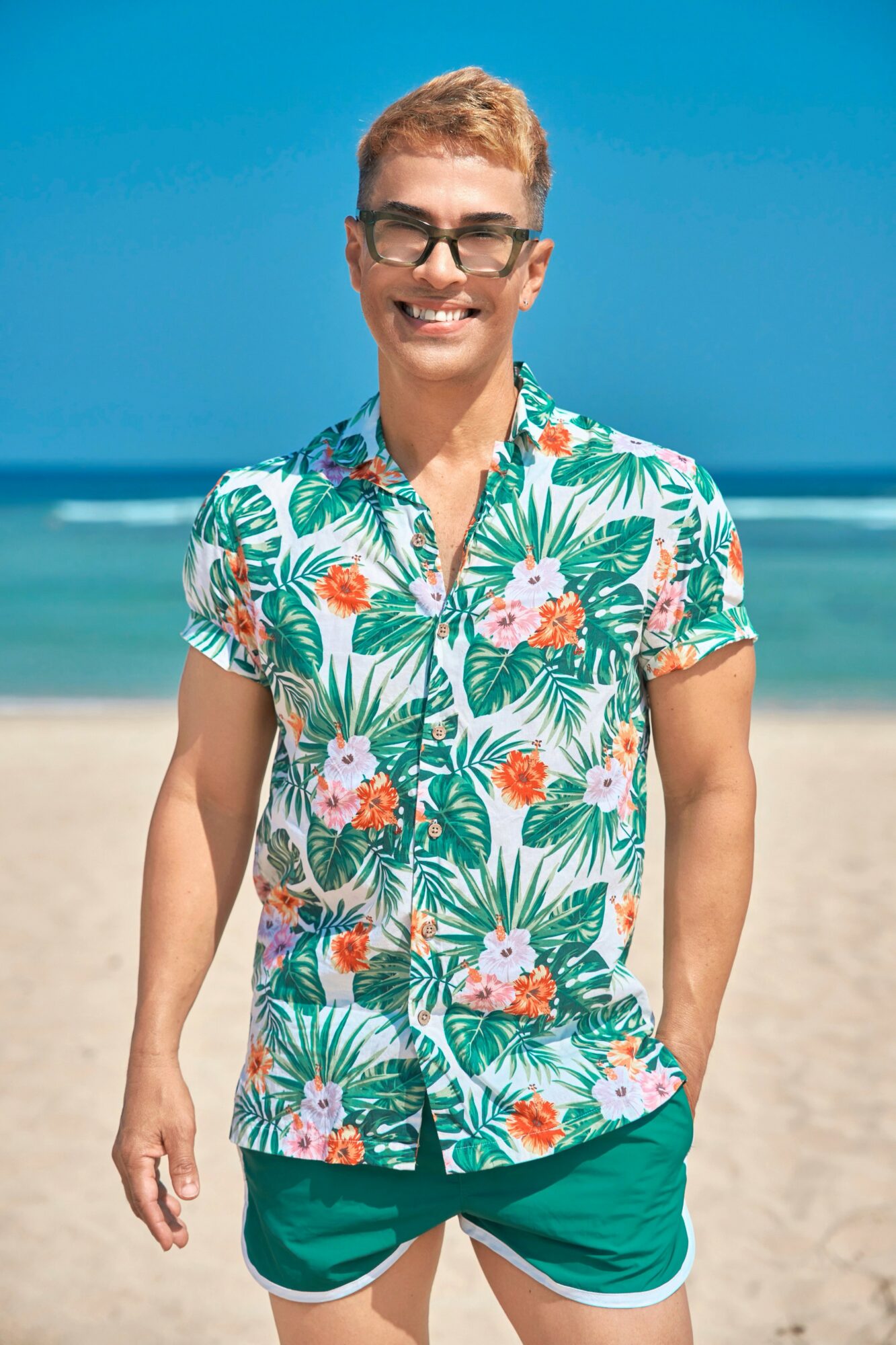 Person smiling on beach wearing a tropical shirt and shorts, with ocean and sky in background.