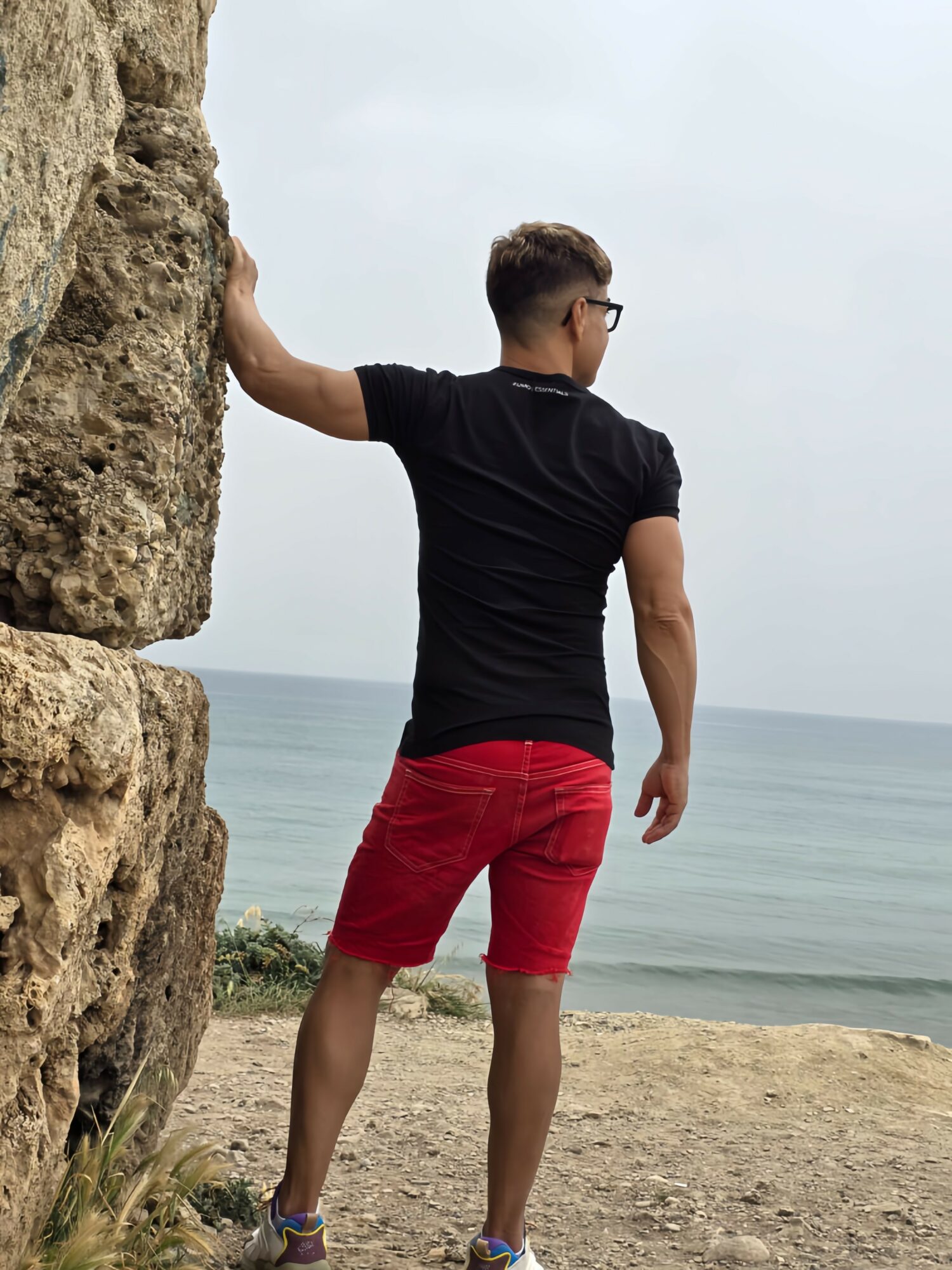 Person standing on a beach, facing the ocean, with one hand on a large rock, wearing a black shirt and red shorts.