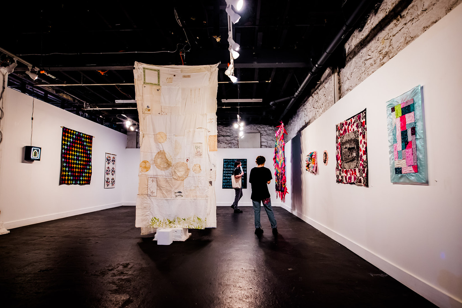 Gallery with colorful abstract paintings on white walls and a large hanging textile art piece in the center, visitors viewing artwork.