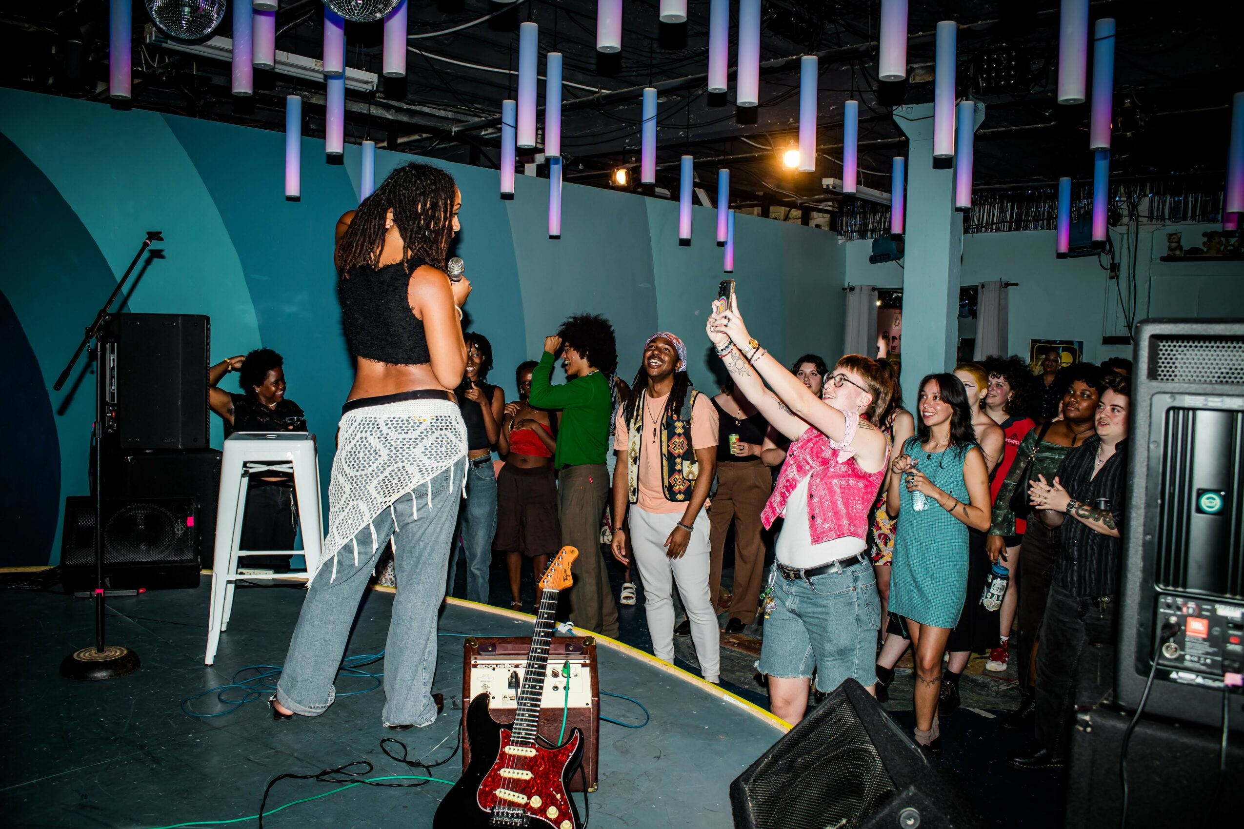 A woman with curly hair stands on stage in front of a group of people in a dimly lit venue with hanging lights, a guitar, and sound equipment.