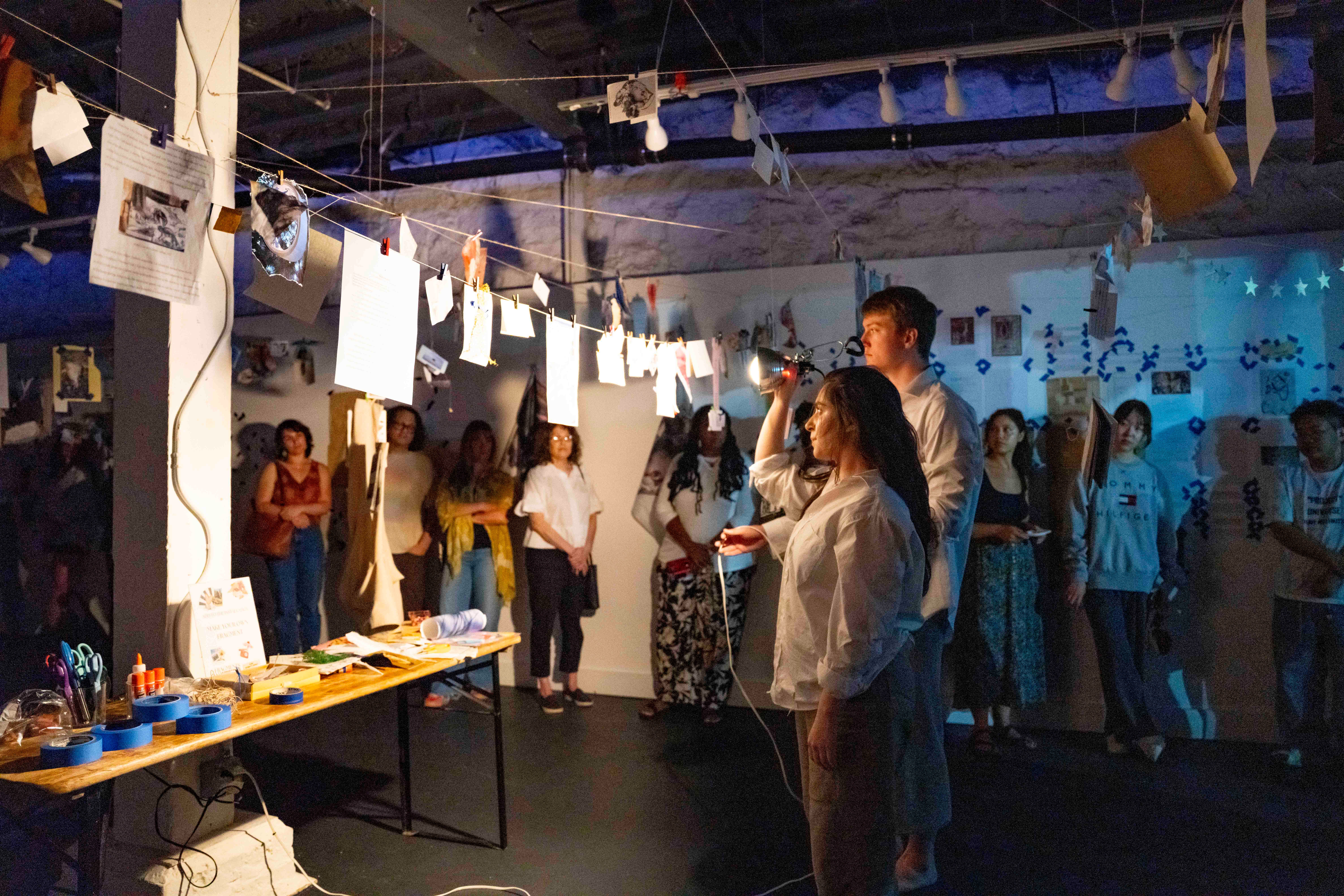 People gather in a room with hanging photos and displays, some looking at tables with items, others talking, blue lighting.