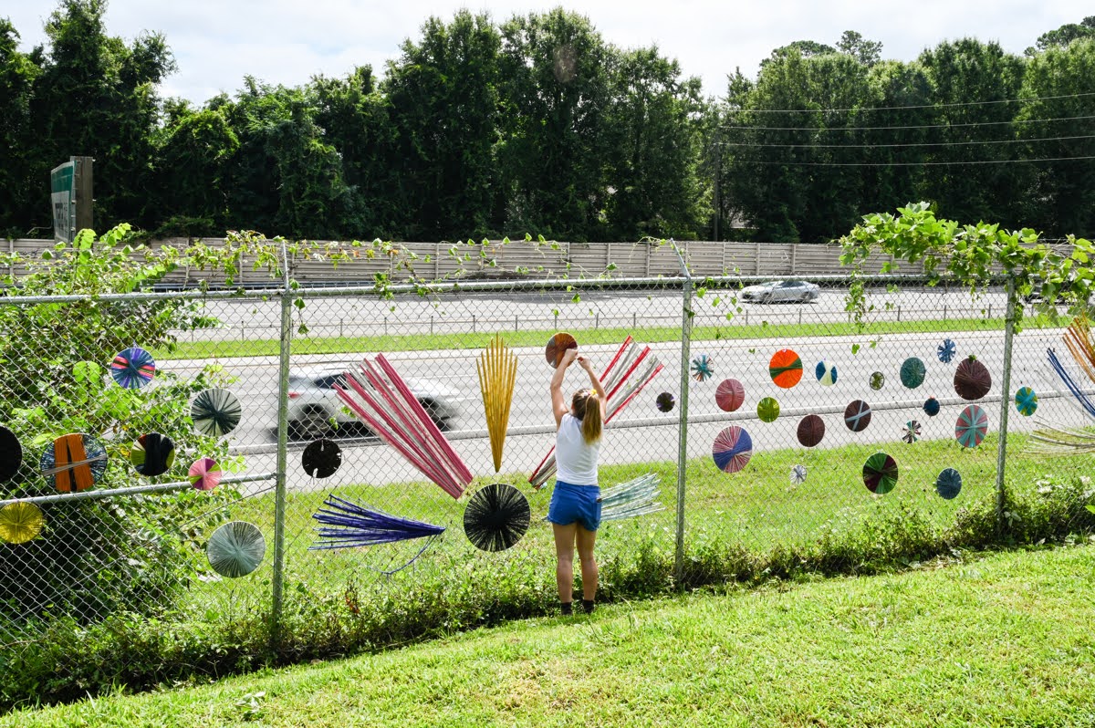 Child playing with colorful balls and objects in a fenced outdoor area with trees in the background.