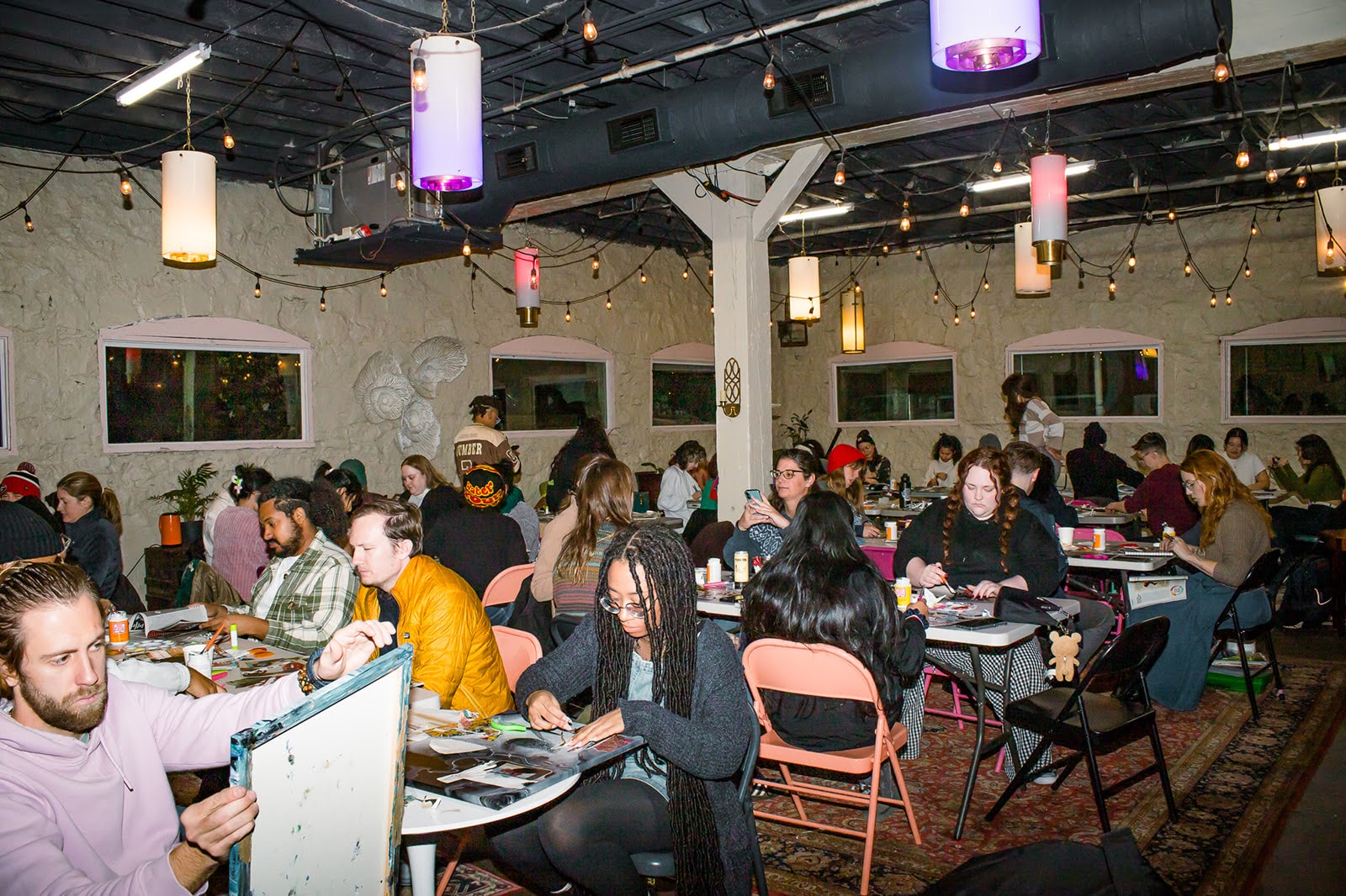 People seated at tables in a decorated indoor space with hanging lights and windows, dining and socializing.