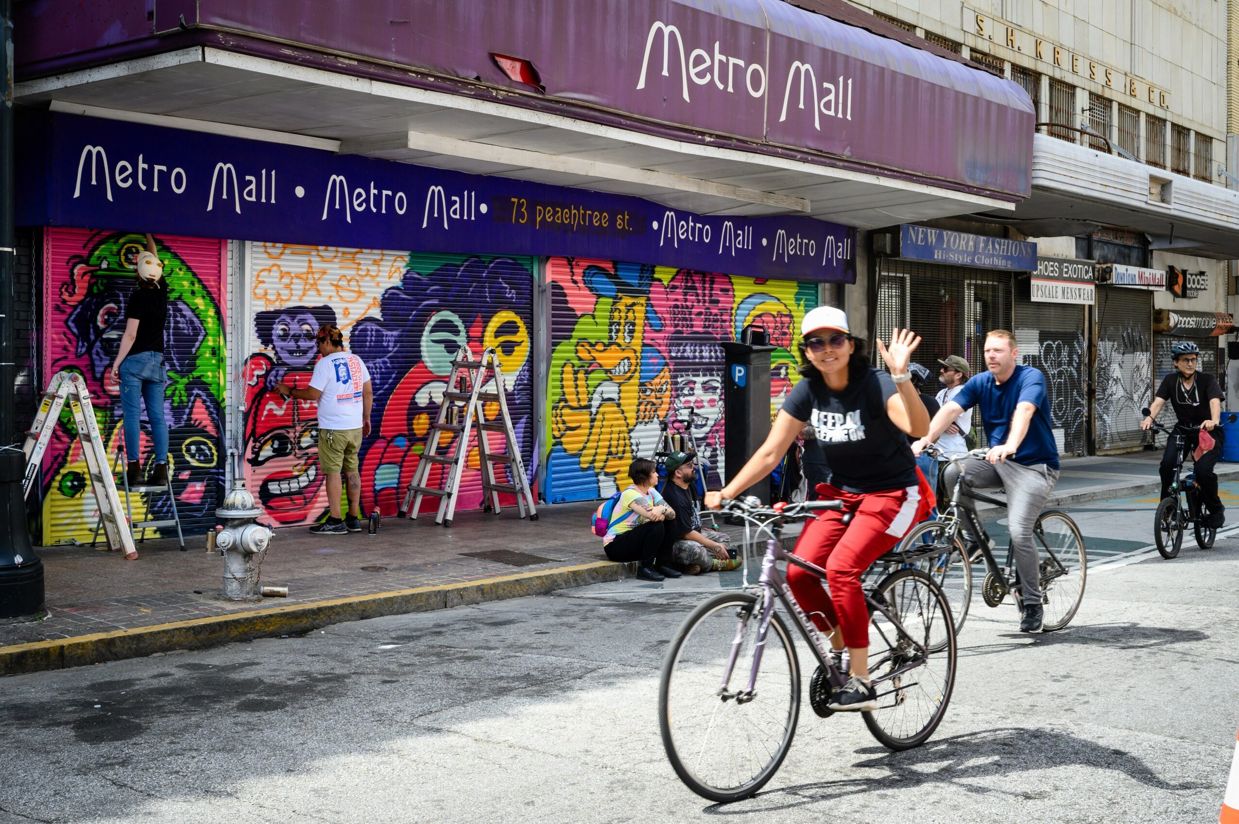 People riding bicycles and walking past a colorful graffiti mural on a city street, with a Metro Mall sign above.