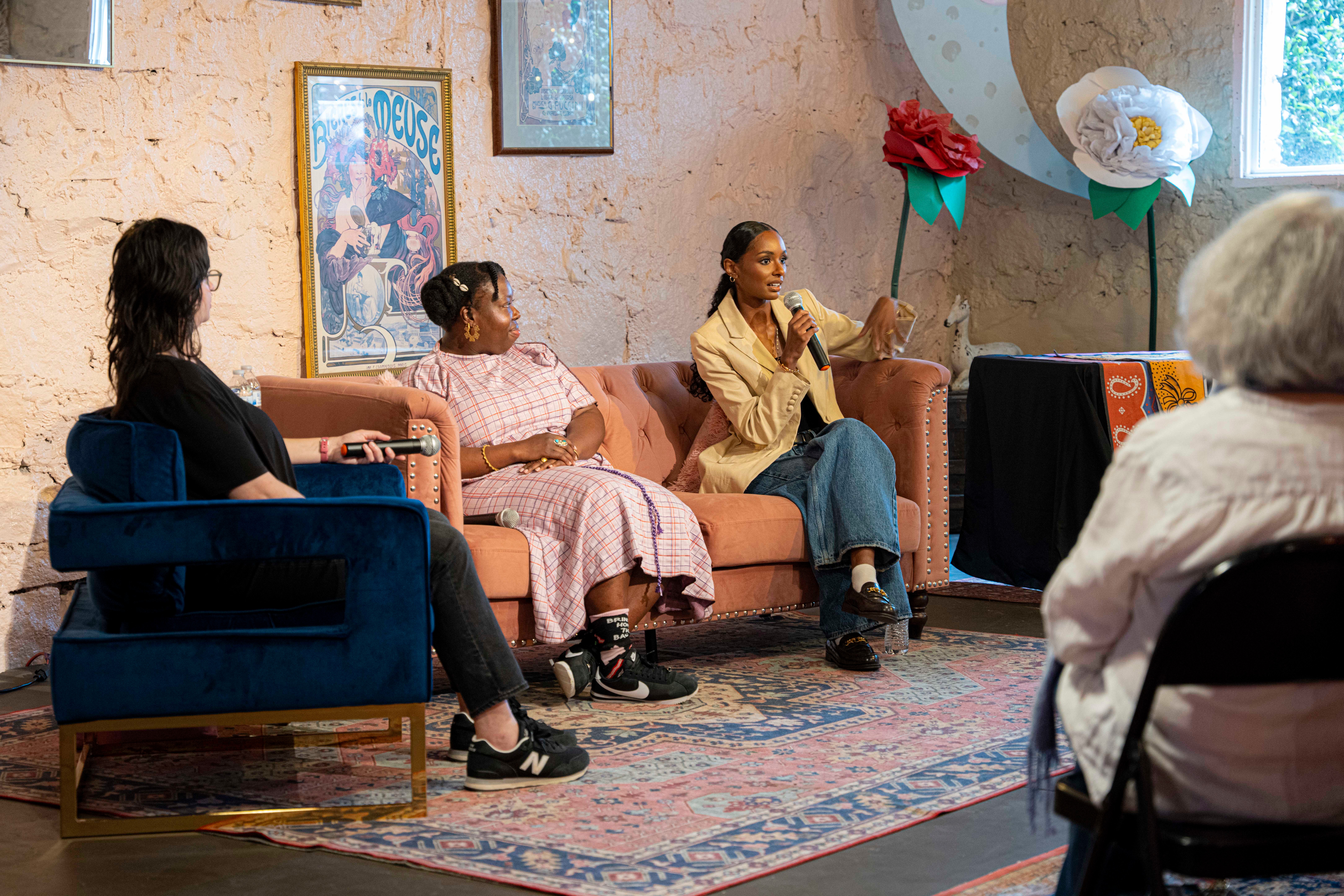 Four women sitting in a room with colorful wall decorations, engaged in conversation, with a patterned rug on the floor.
