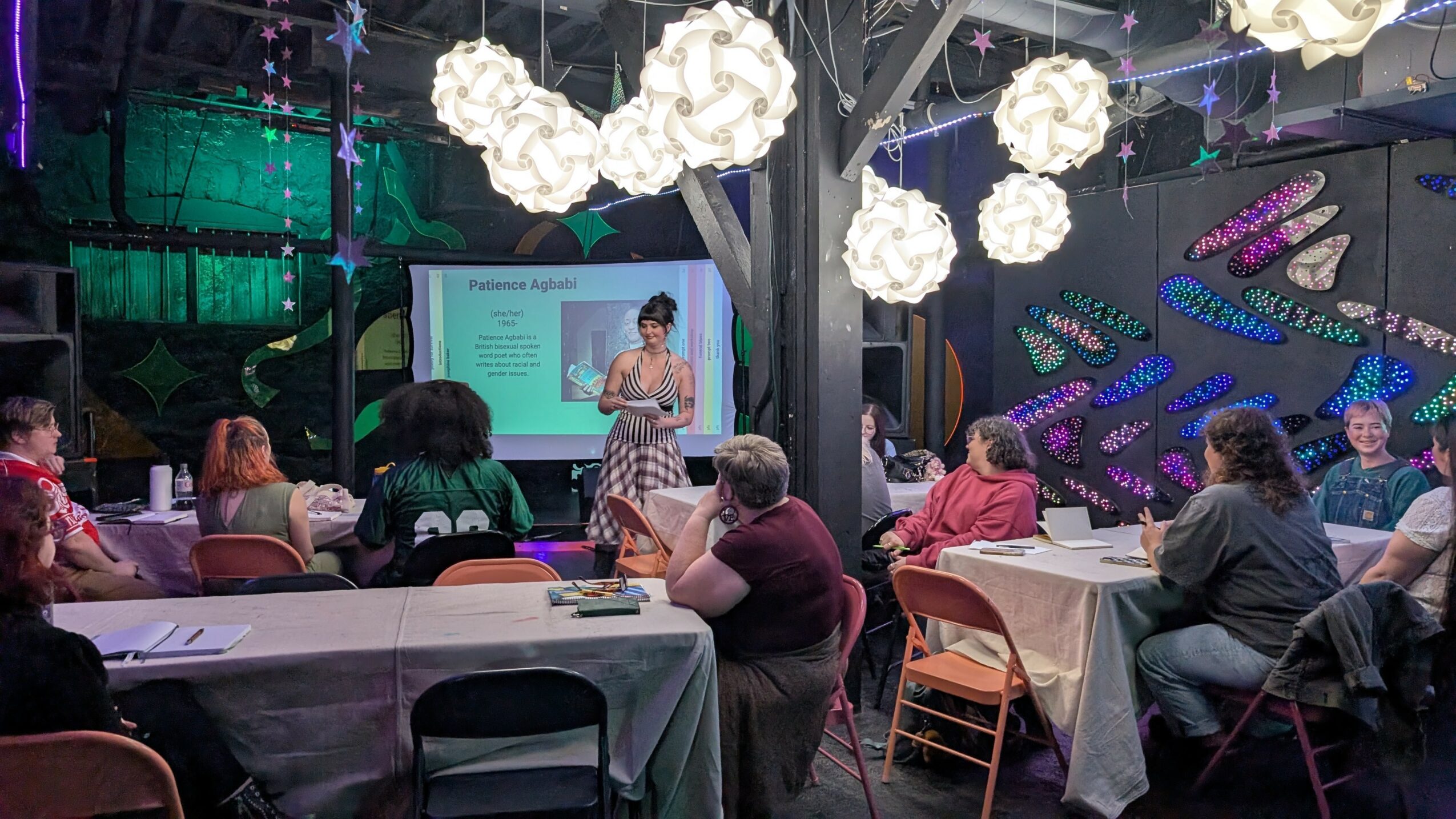 A woman presents to an audience in a dimly lit room with decorative hanging lights and colorful wall art.