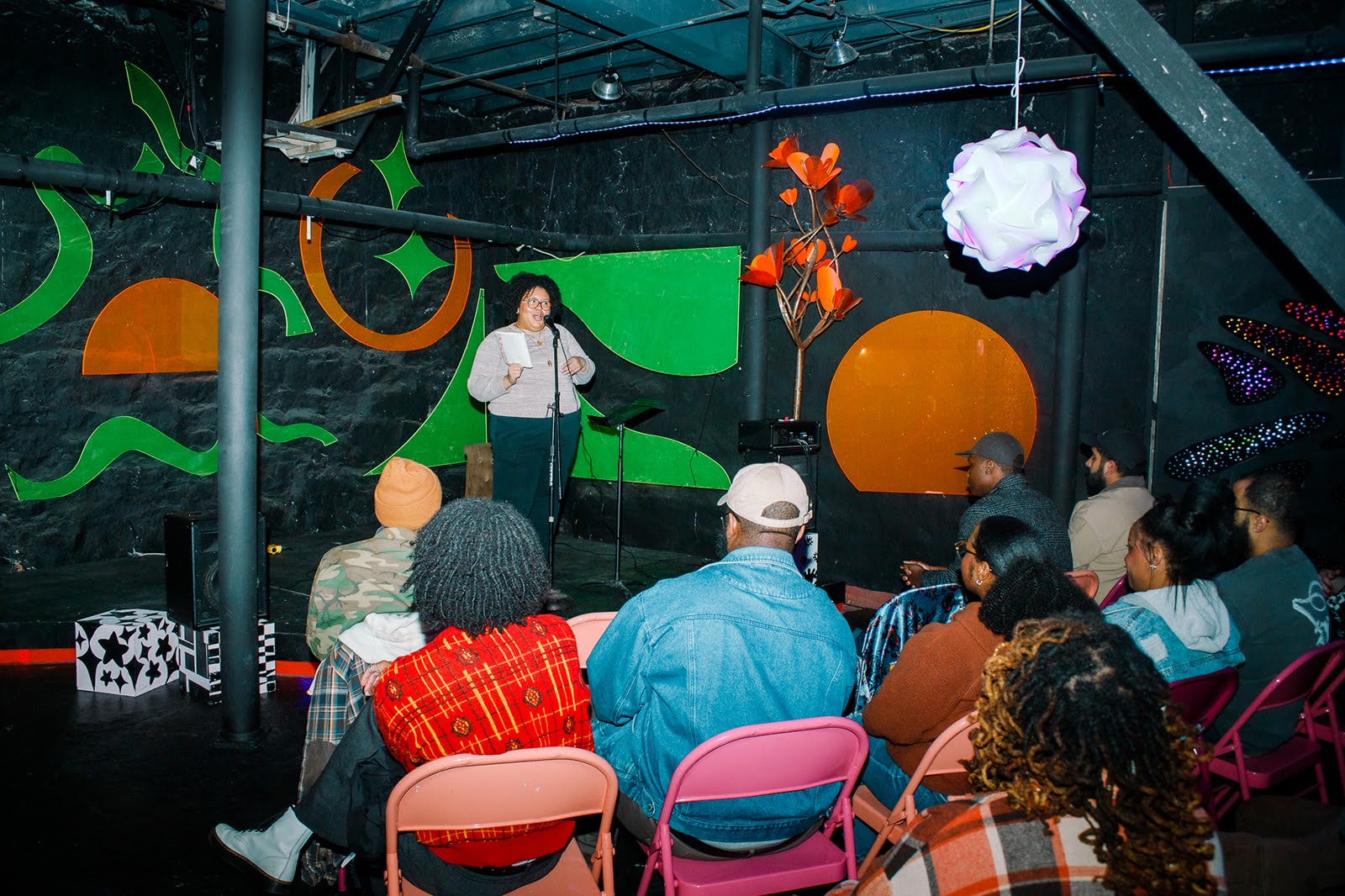 Person speaking on stage with audience seated in chairs, colorful abstract background, stage lighting, and decorations.