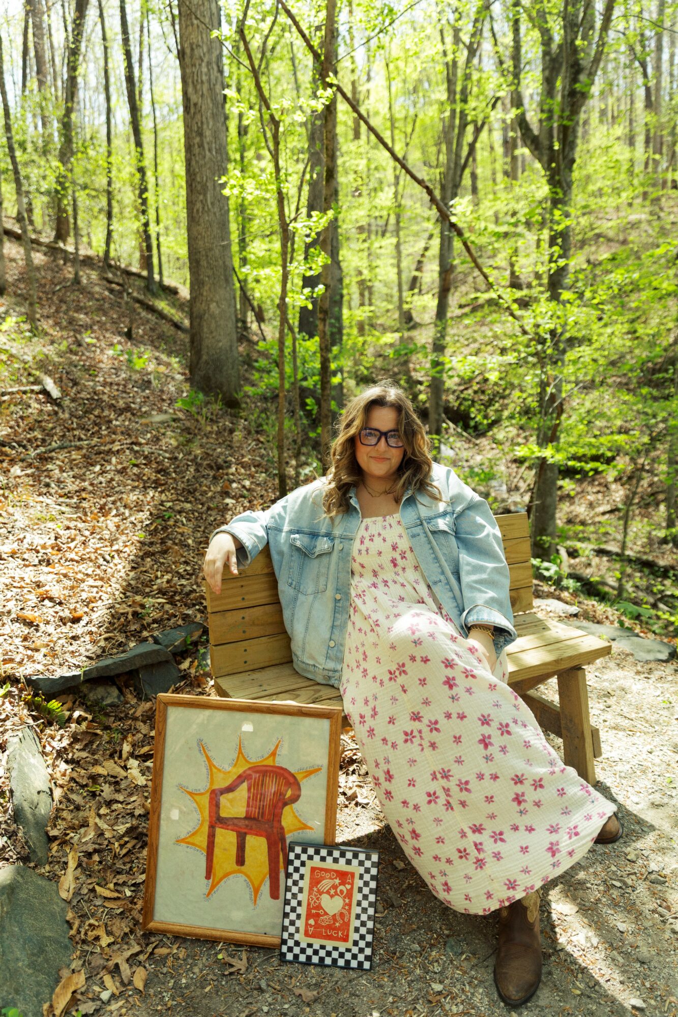 Woman sitting on a bench in a forest, wearing glasses and a denim jacket, with framed artwork on the ground.