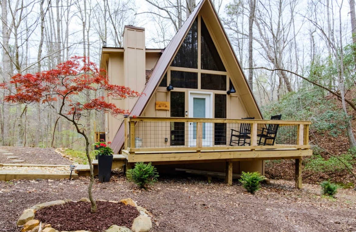 A small A-frame house with a balcony, surrounded by trees and a landscaped yard, during daytime.
