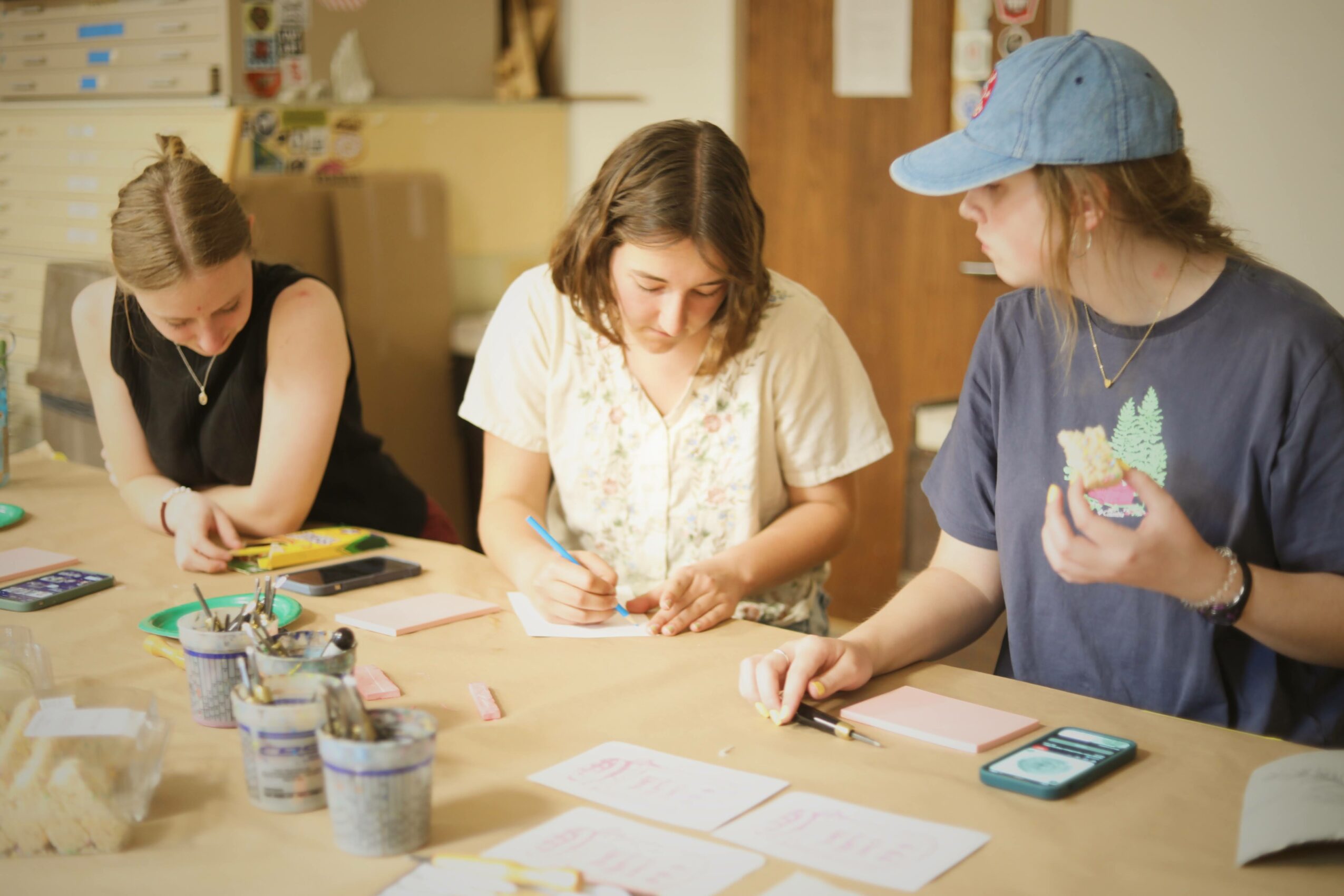 Three women sit at a table working on a project with papers, pens, and containers of supplies, in a room with shelves.