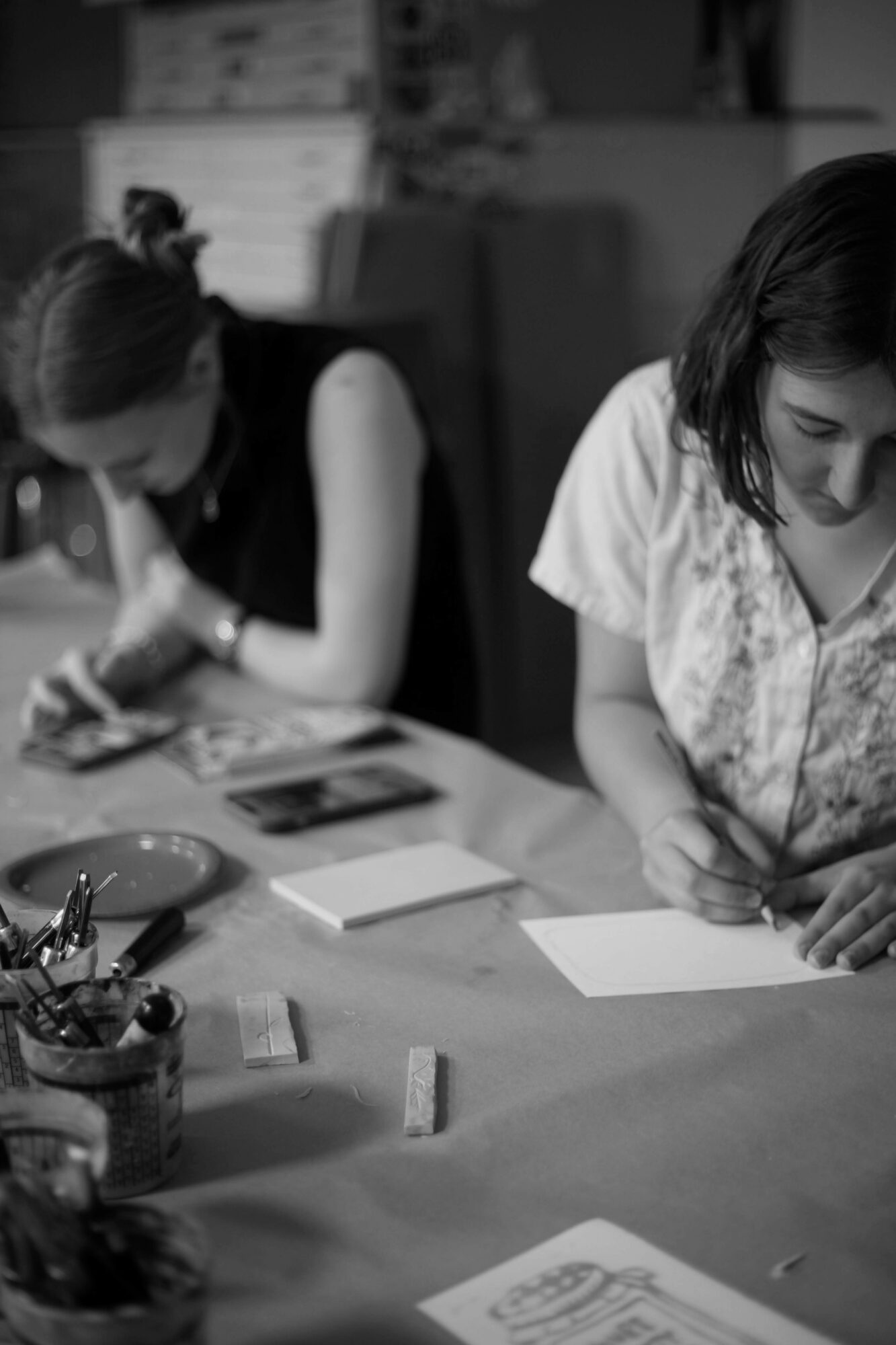 Two women sitting at a table, writing on paper, with art supplies and papers nearby.